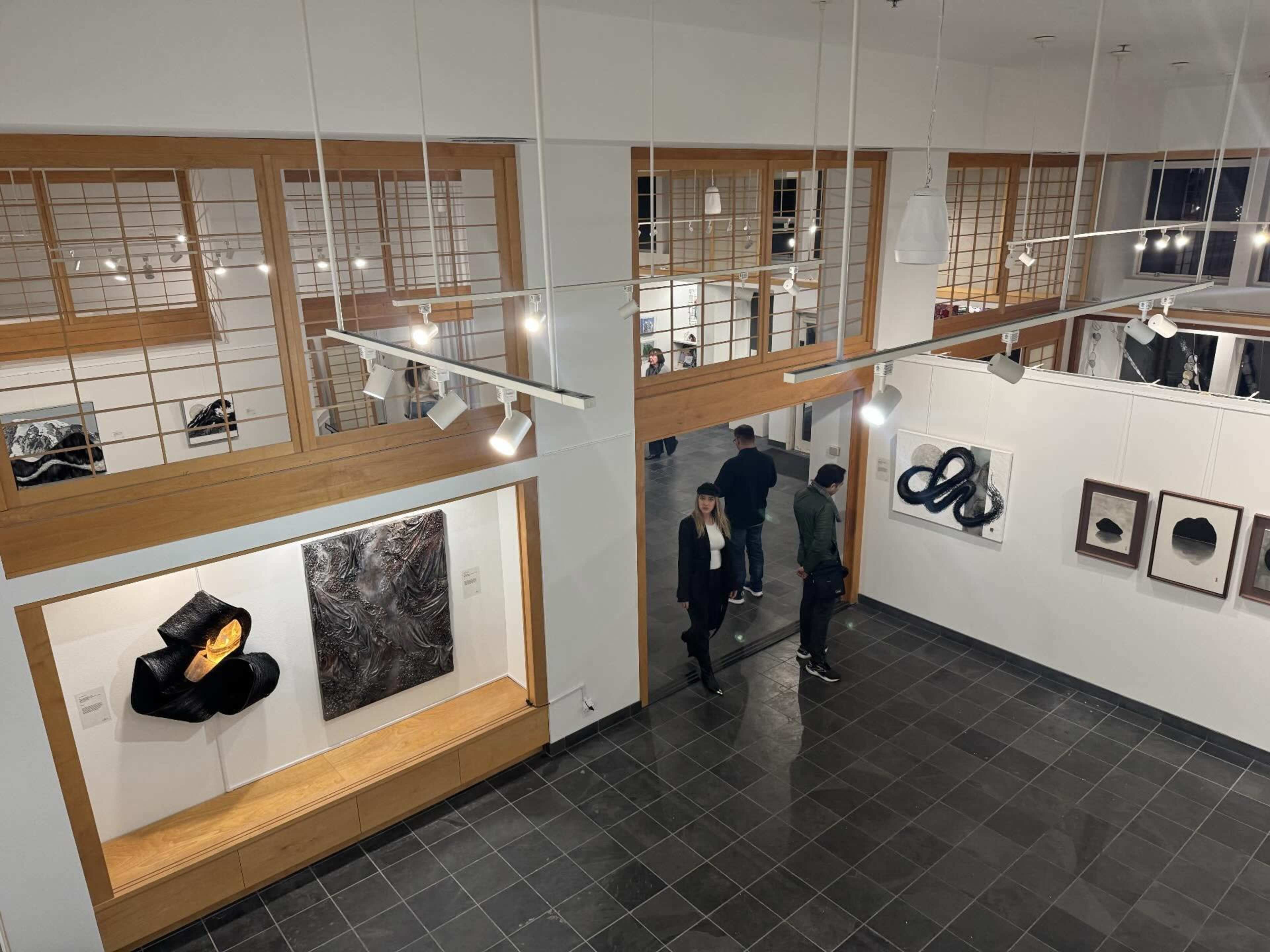 The image depicts an art gallery with wooden accents, featuring artworks on the walls and visitors walking through the space.