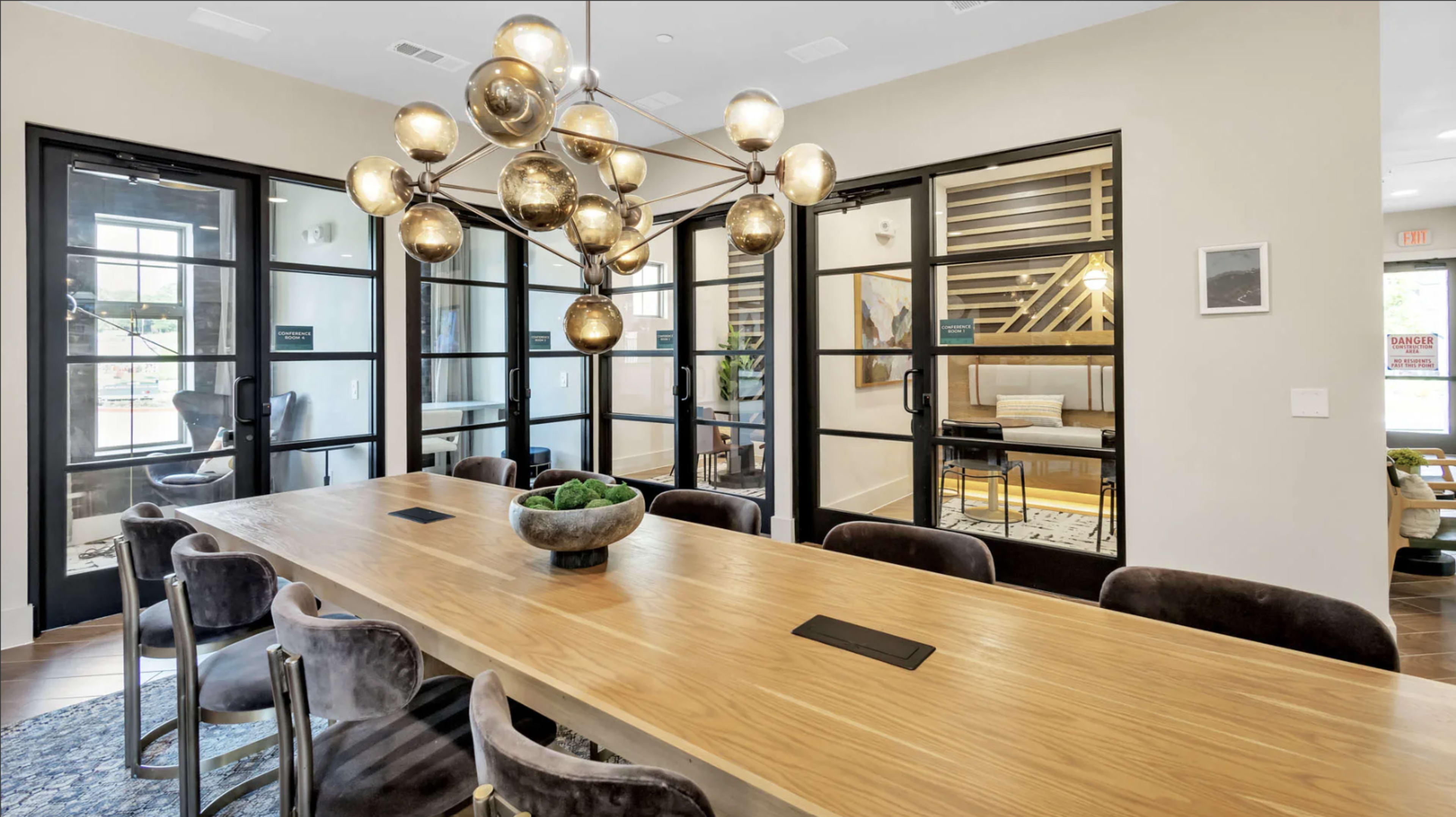 A large wooden table with dark chairs is surrounded by glass doors and a modern chandelier in a well-lit meeting space.
