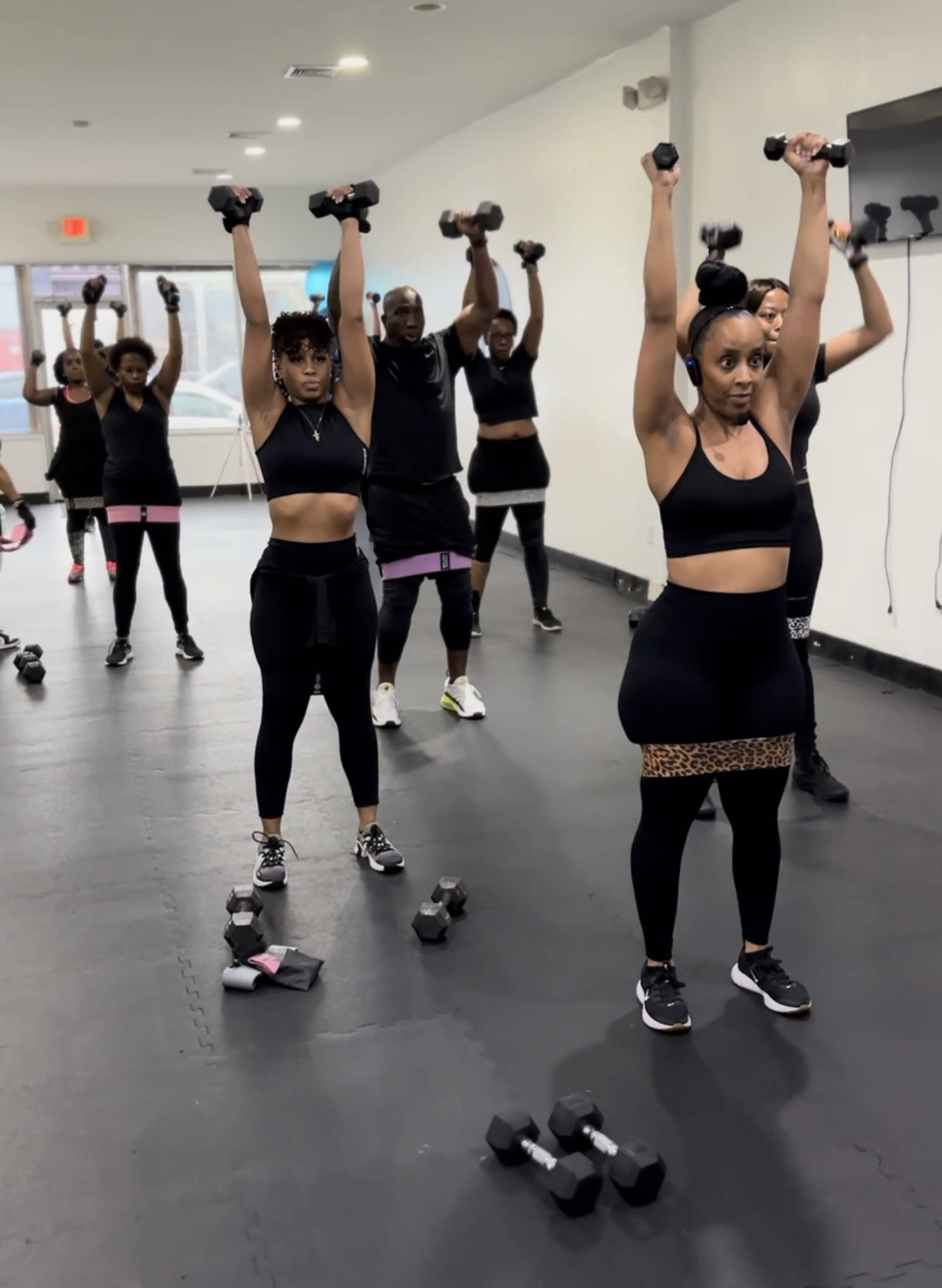A group of individuals in black workout attire are performing weightlifting exercises with dumbbells in a gym.