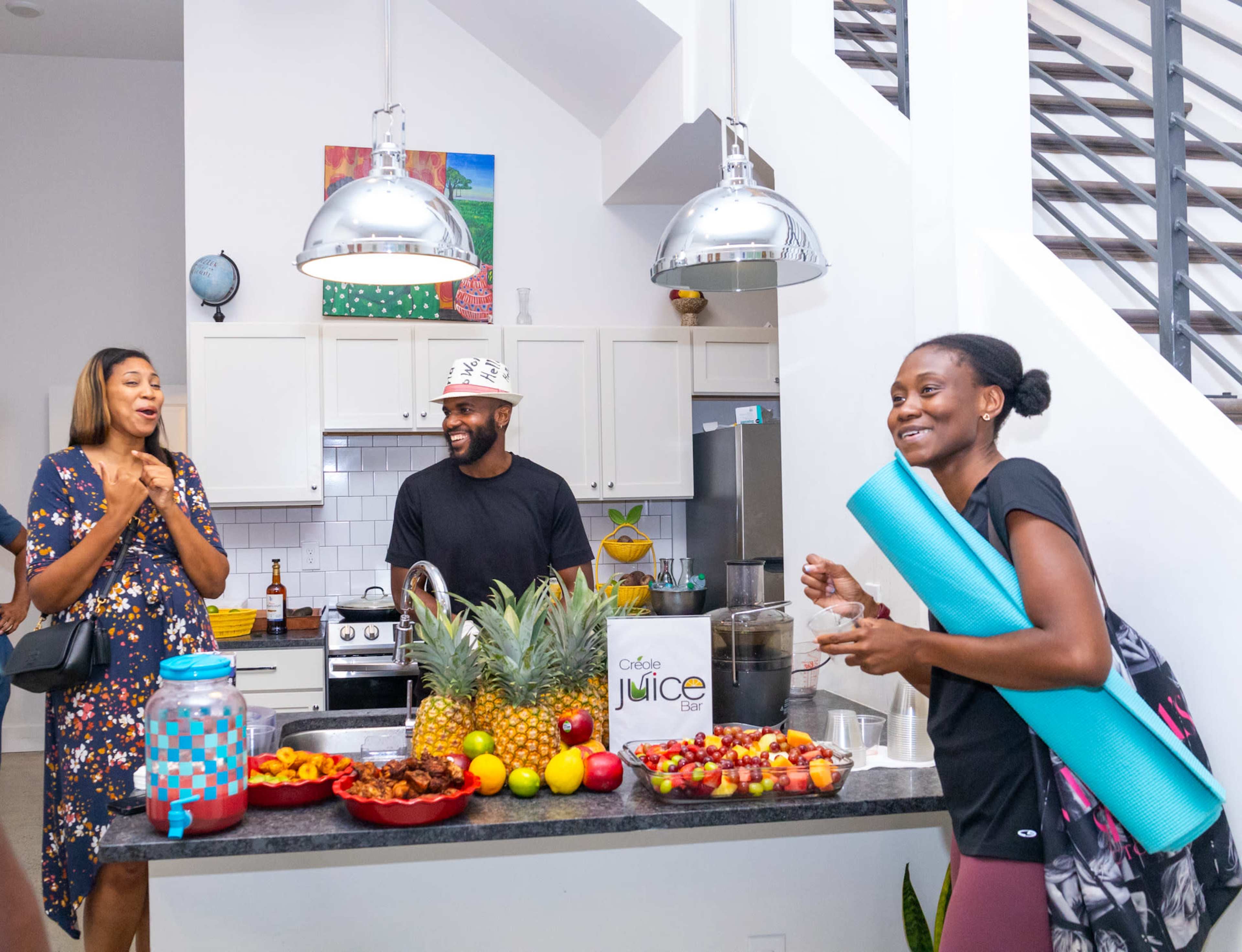 A group of three people stands around a kitchen island filled with fresh fruits and a juice maker, smiling and engaging in conversation.