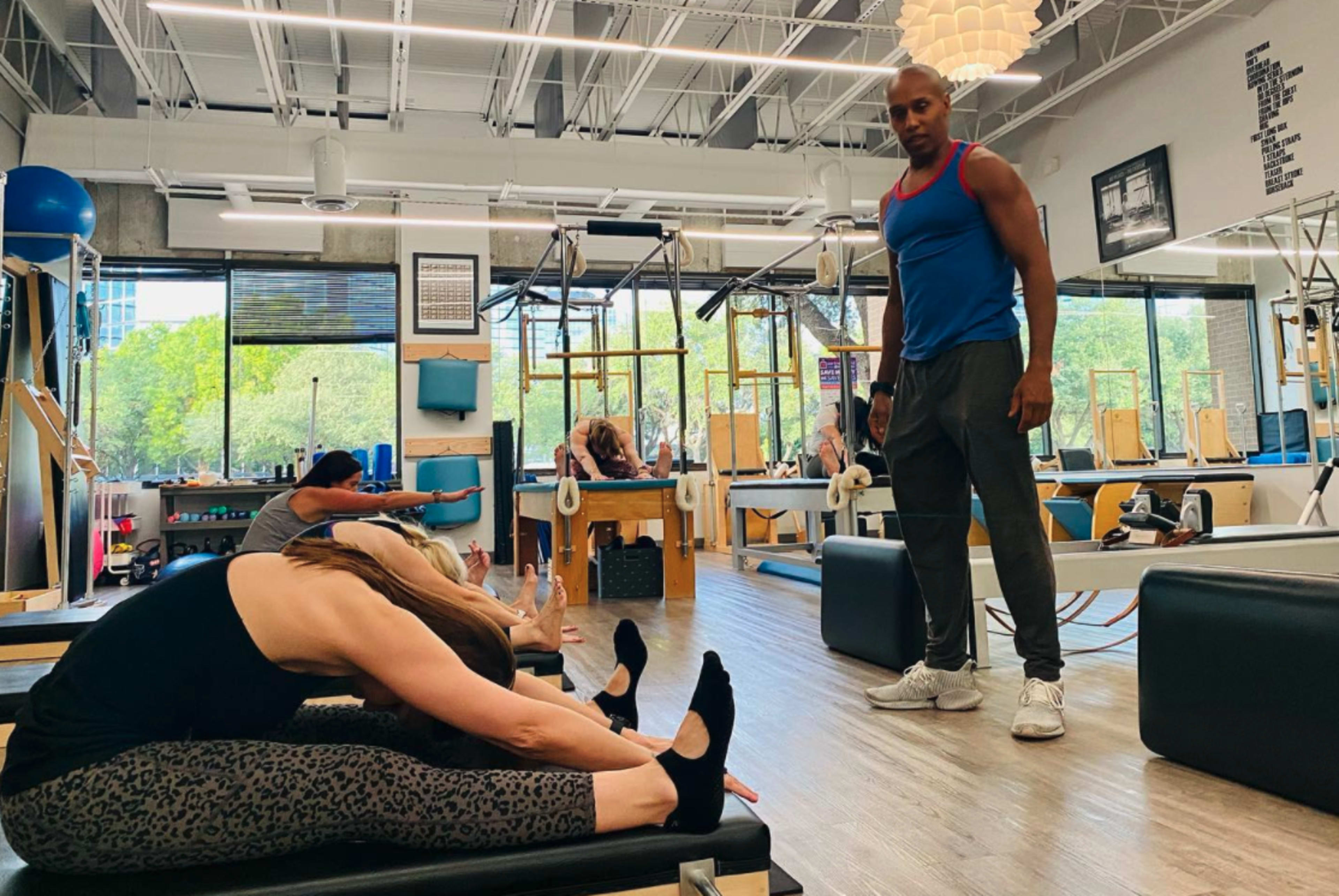 A fitness instructor supervises students performing stretching exercises in a Pilates studio.
