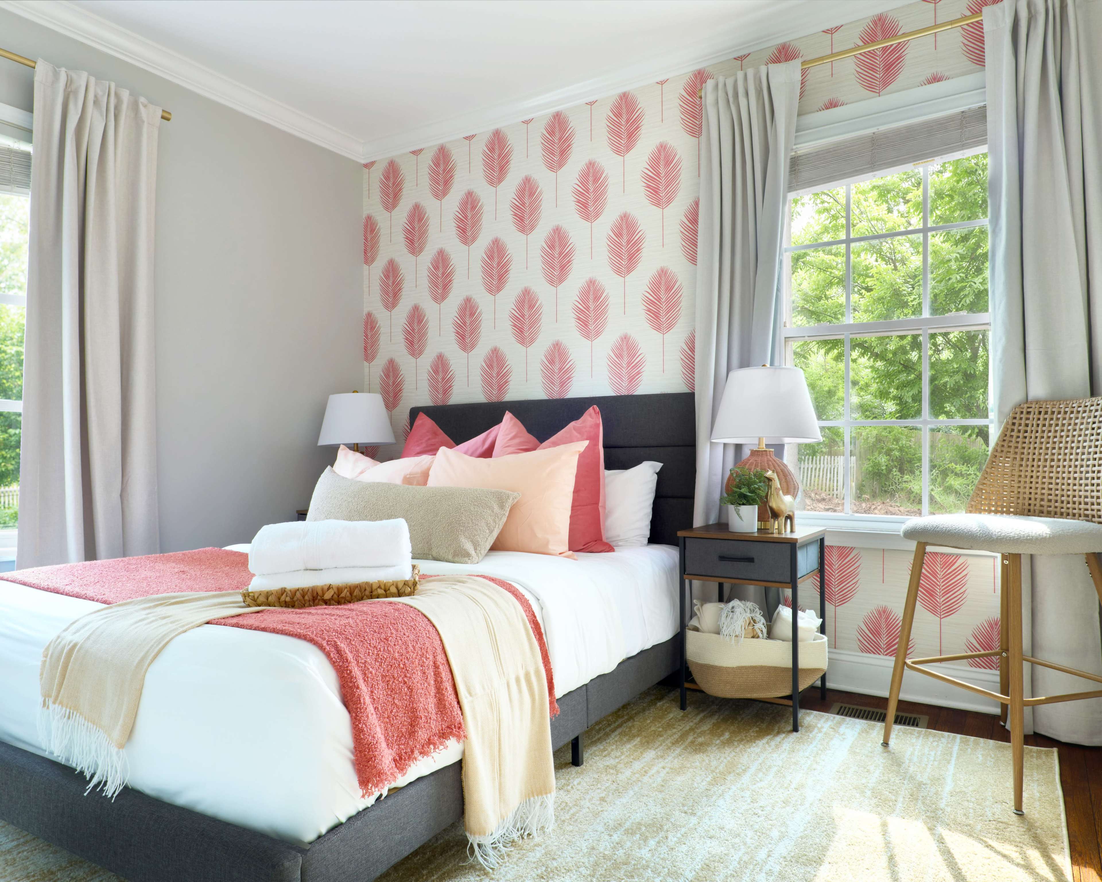 The image shows a well-decorated bedroom featuring a gray and pink leaf-patterned accent wall, a bed with pastel-colored pillows and a blanket, and a wooden nightstand beside it.