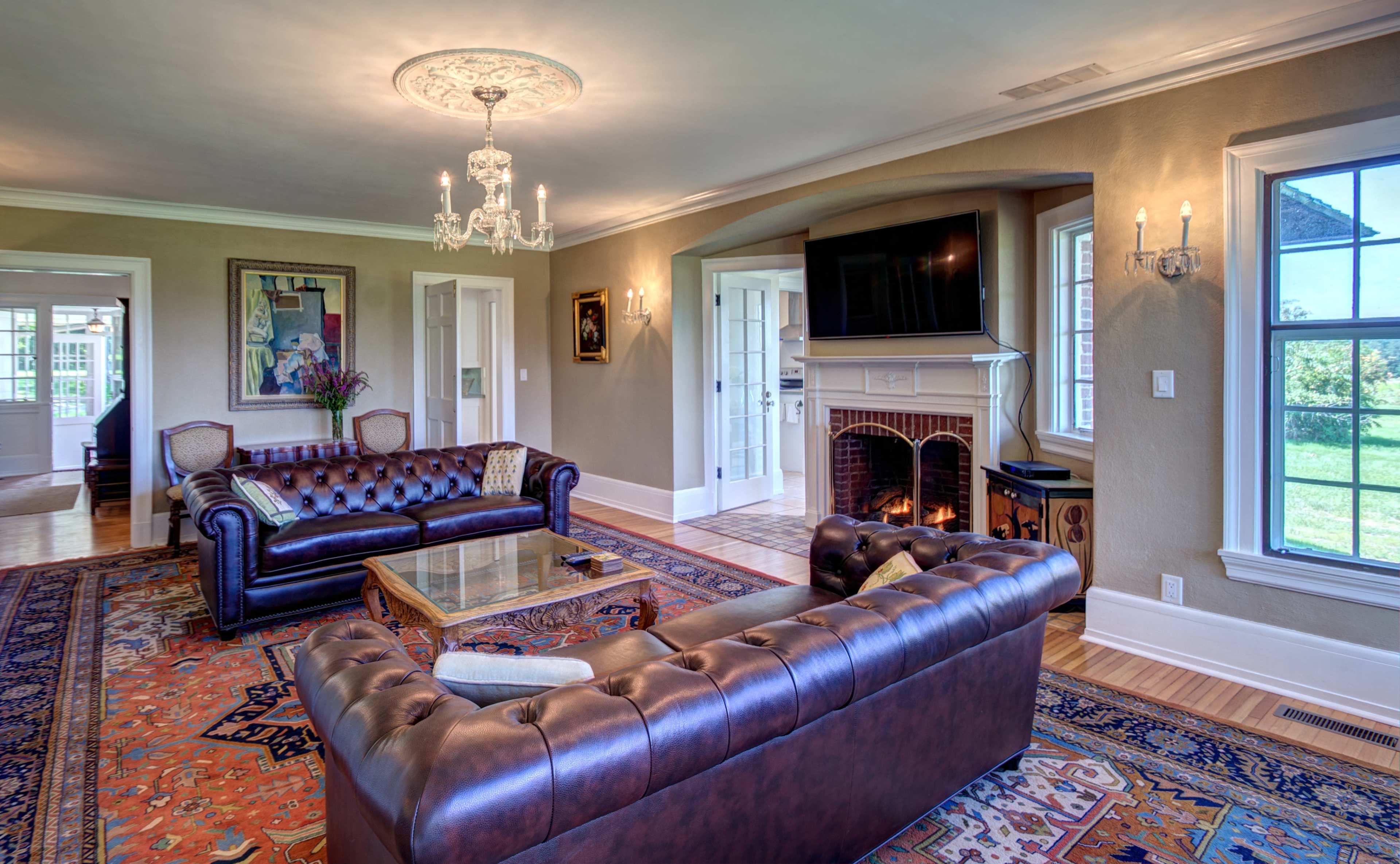 A spacious living room with a leather sofa, armchairs, a coffee table, a fireplace, and large windows overlooking a garden.