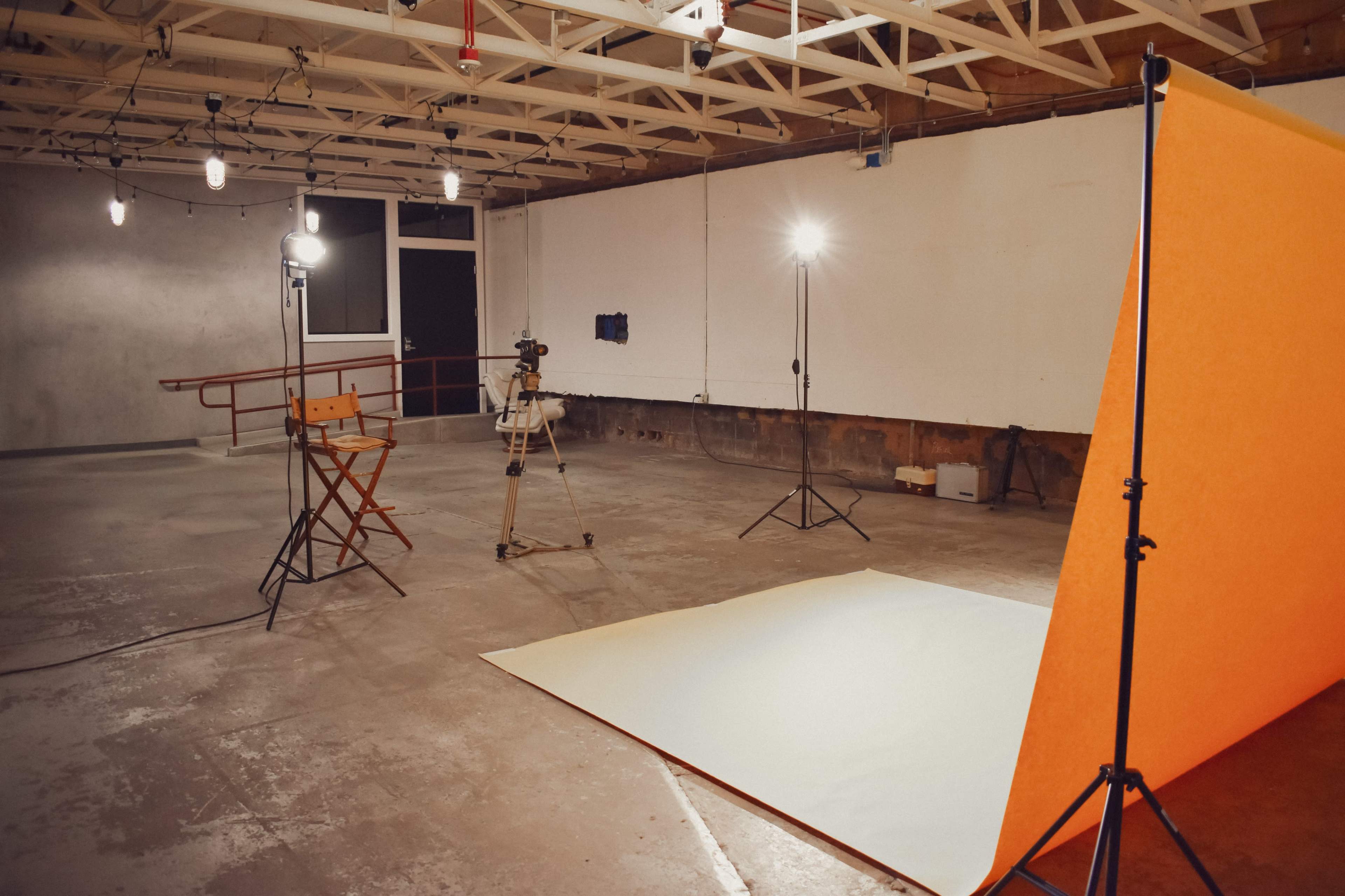 A photography studio is set up with a large orange backdrop, a director's chair, and several lighting equipment positioned around a concrete floor.