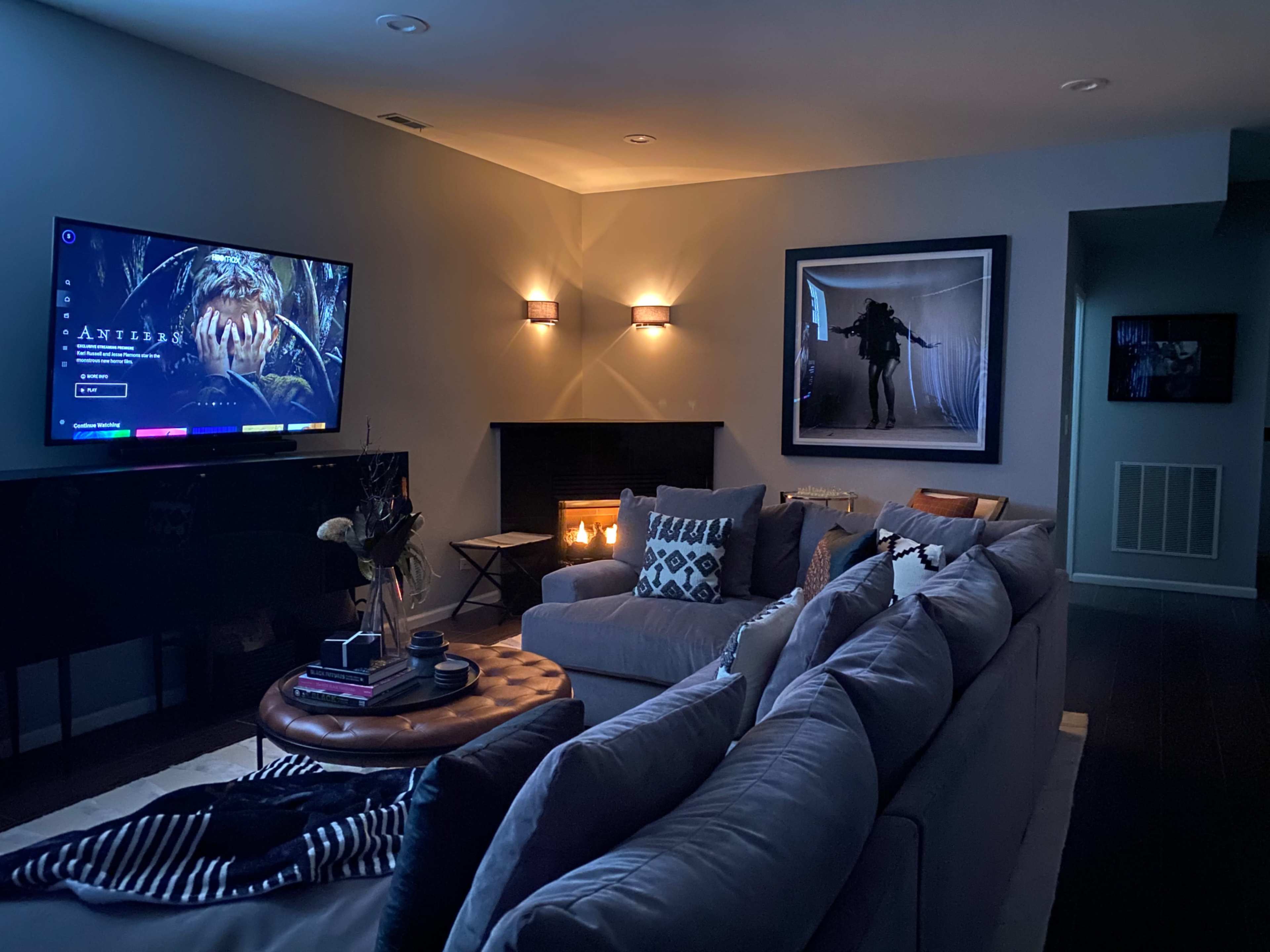 A cozy living room features a large television displaying a digital interface, a sectional sofa, a round coffee table, and wall-mounted lights illuminating the space.
