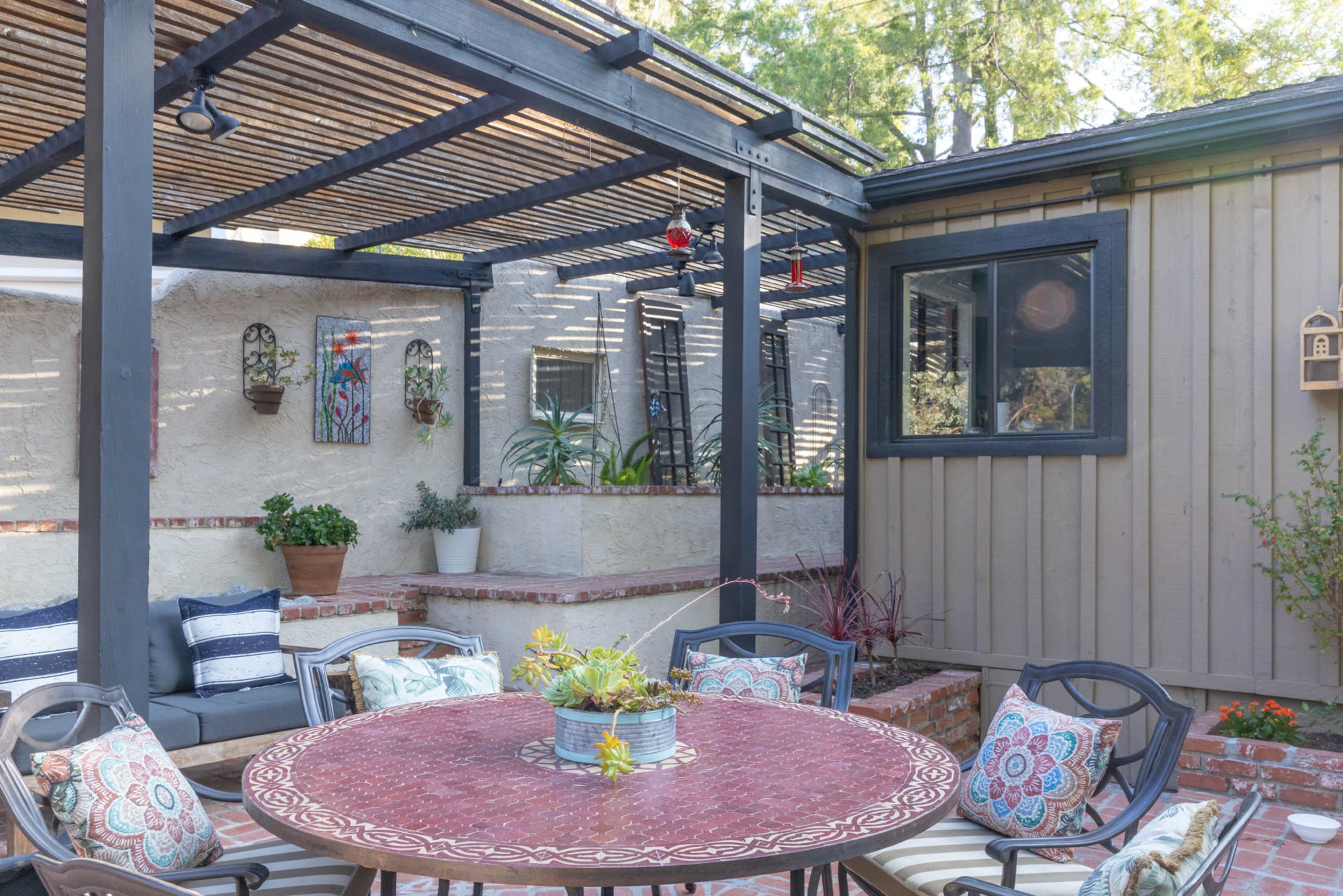 A patio area features a circular table surrounded by chairs, with a shaded section supported by a wooden trellis and adjacent to a dwelling.