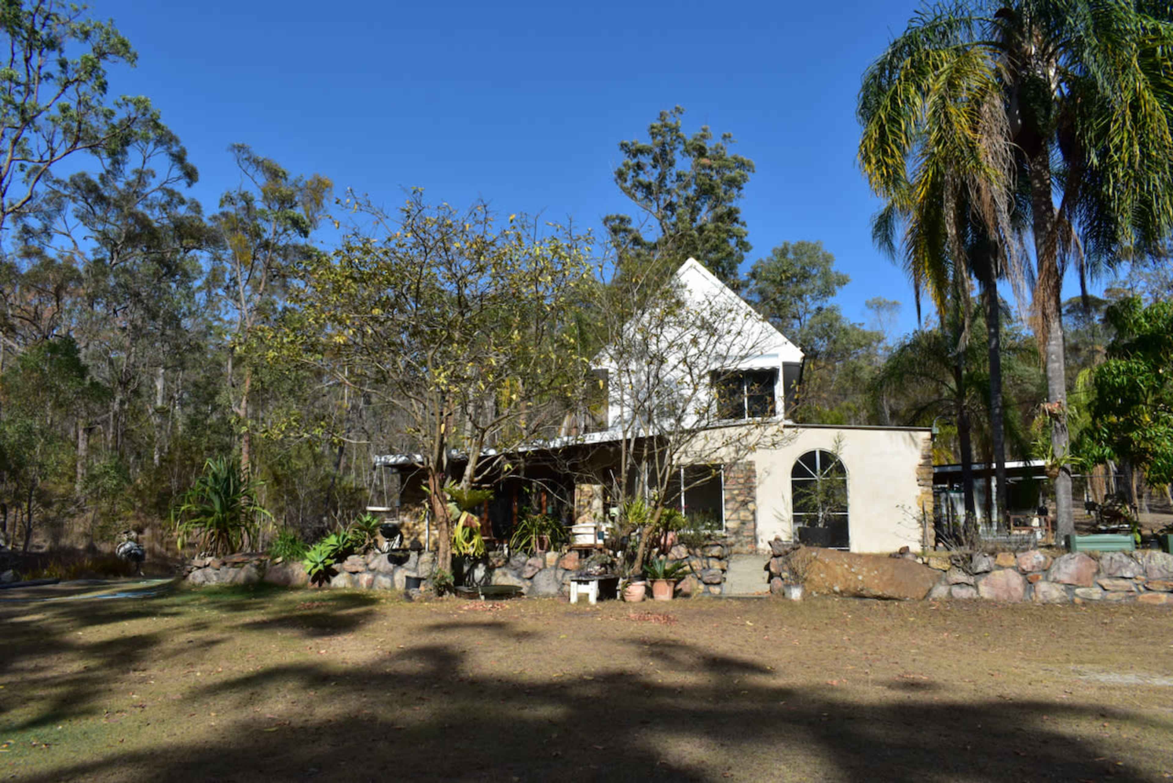 A two-story house with a triangular roof is surrounded by trees and landscaped rocks in a rural outdoor setting.