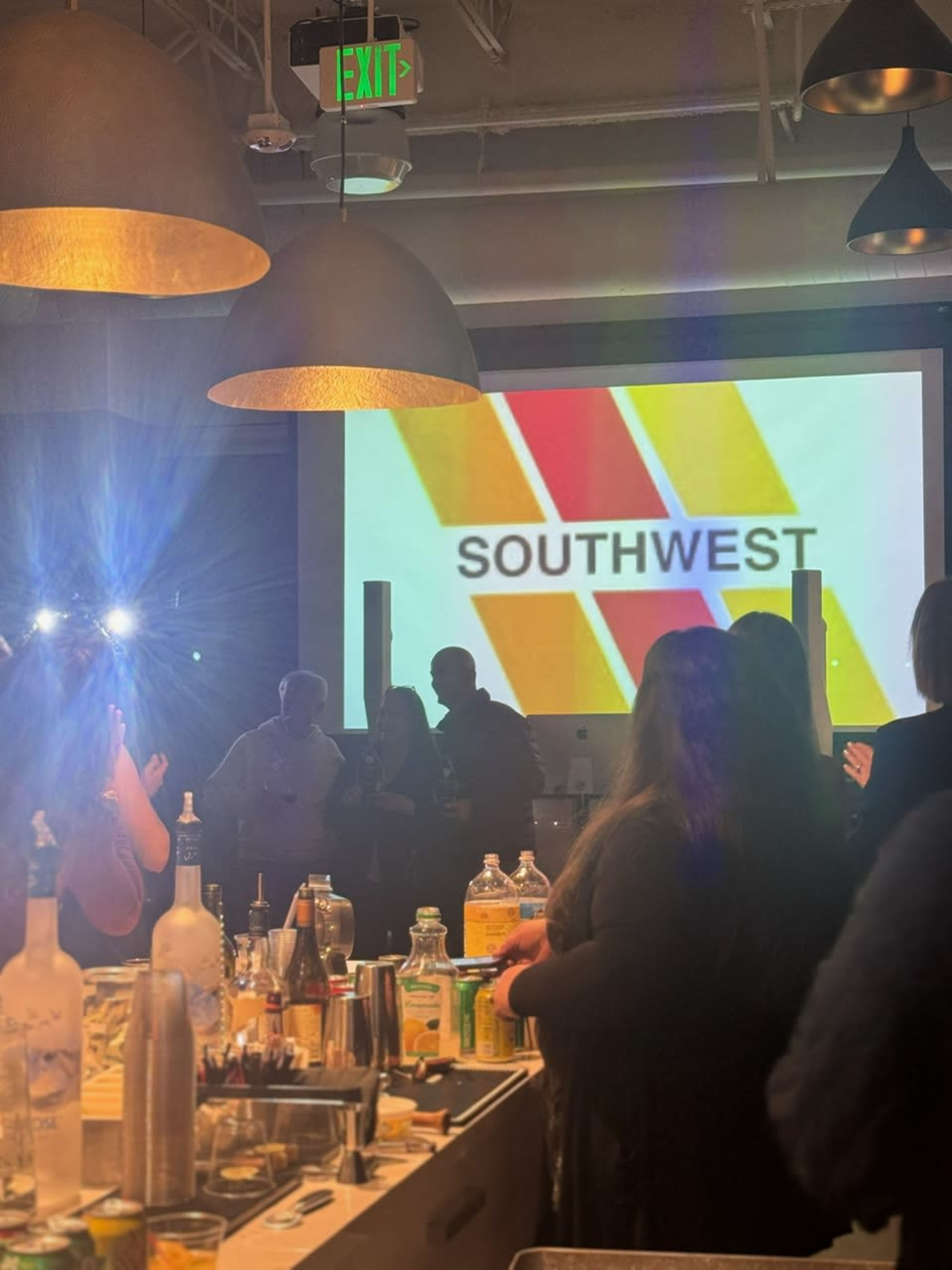 A crowd gathers around a bar as a presentation displaying the "SOUTHWEST" logo is projected on a screen.