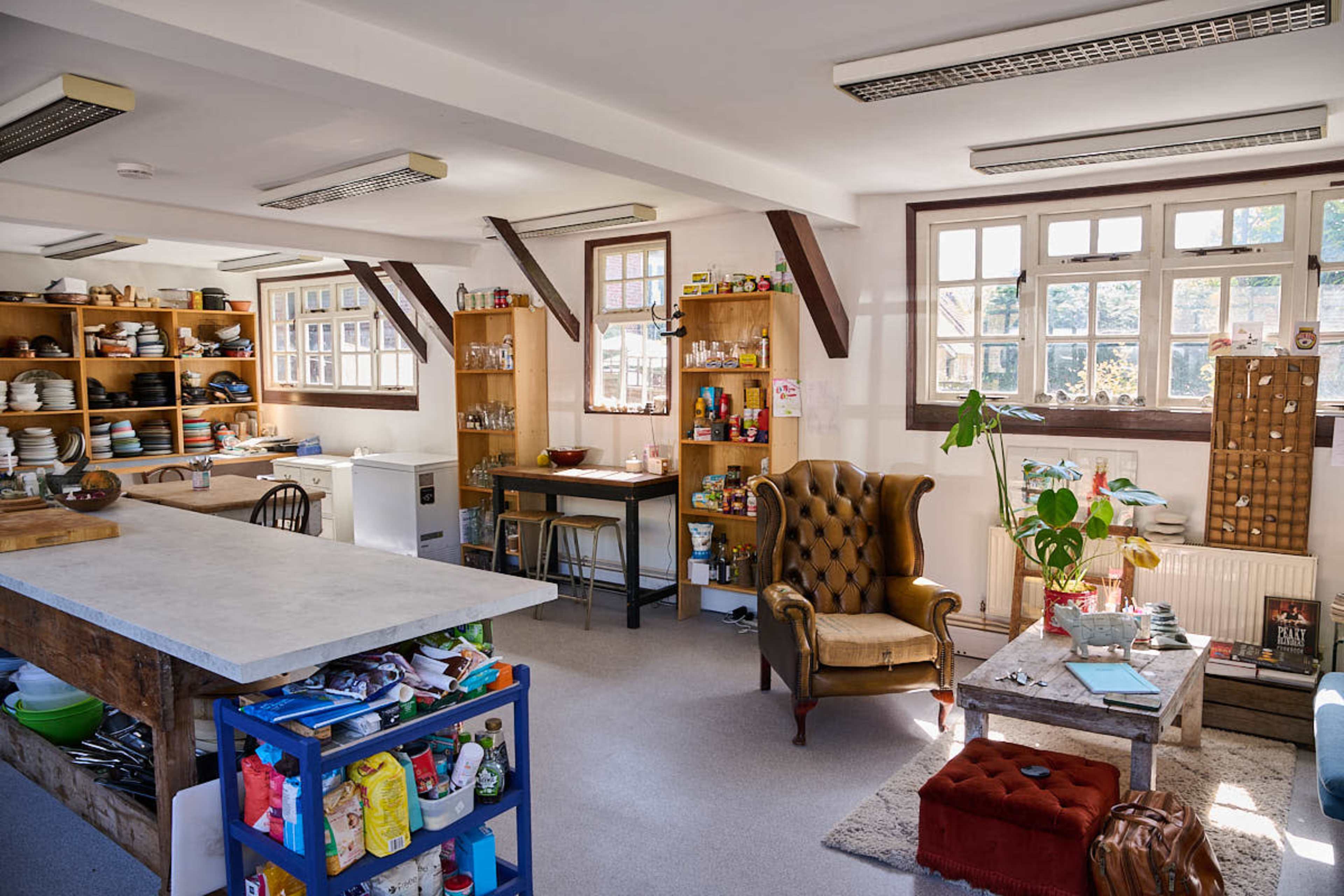 The image shows a spacious kitchen area with wooden shelves filled with dishes and pantry items, a central island, and a cozy seating arrangement including a green armchair and a small coffee table.