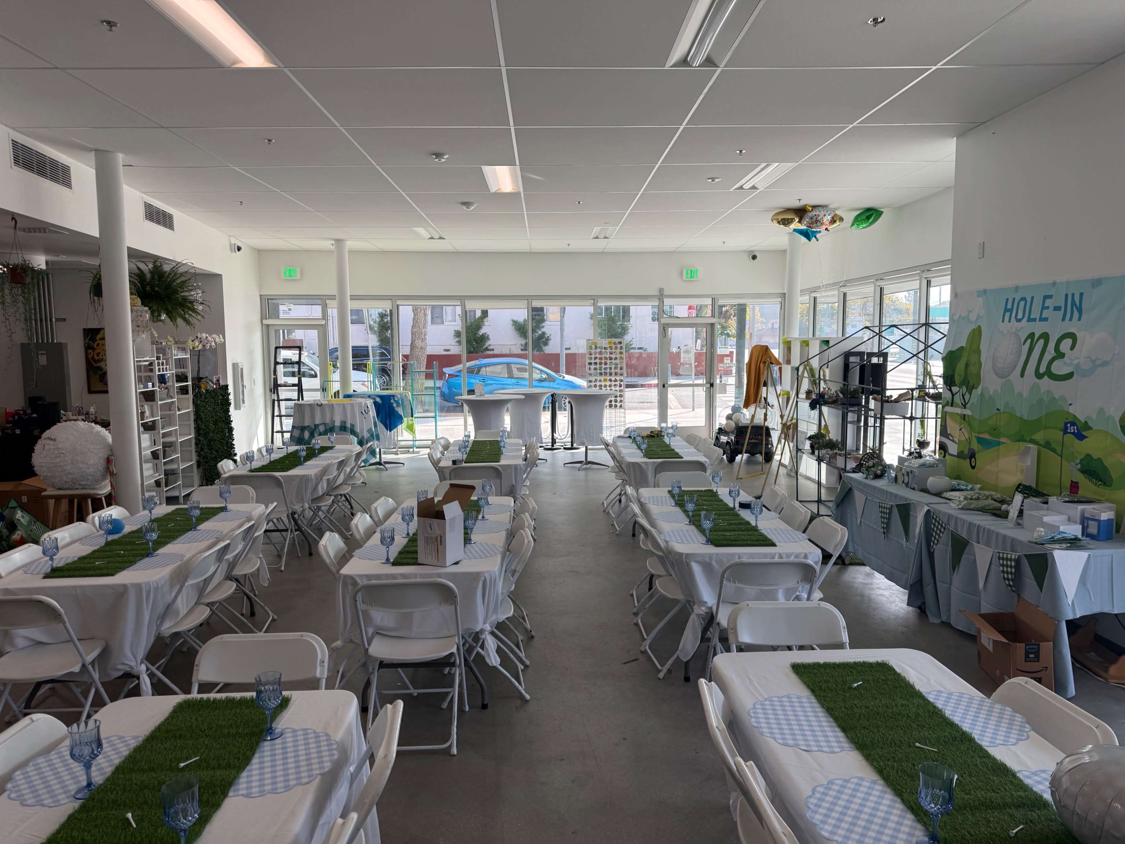 The image shows a spacious event room set up with multiple tables covered in white tablecloths and decorated with green grass-like centerpieces, ready for an occasion.