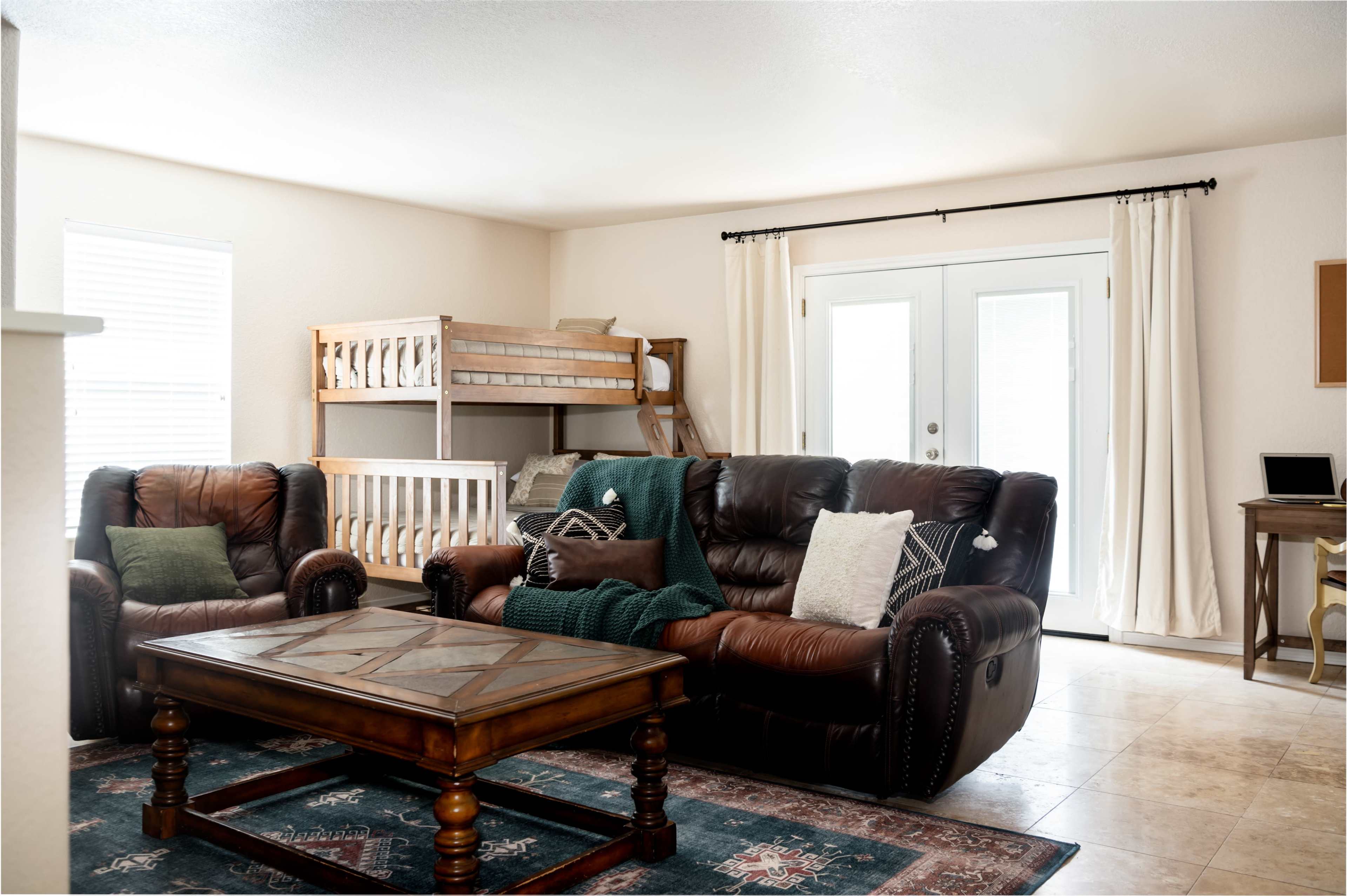 A cozy living room features two leather sofas facing a wooden coffee table, with a bunk bed in the background and sliding glass doors leading outside.