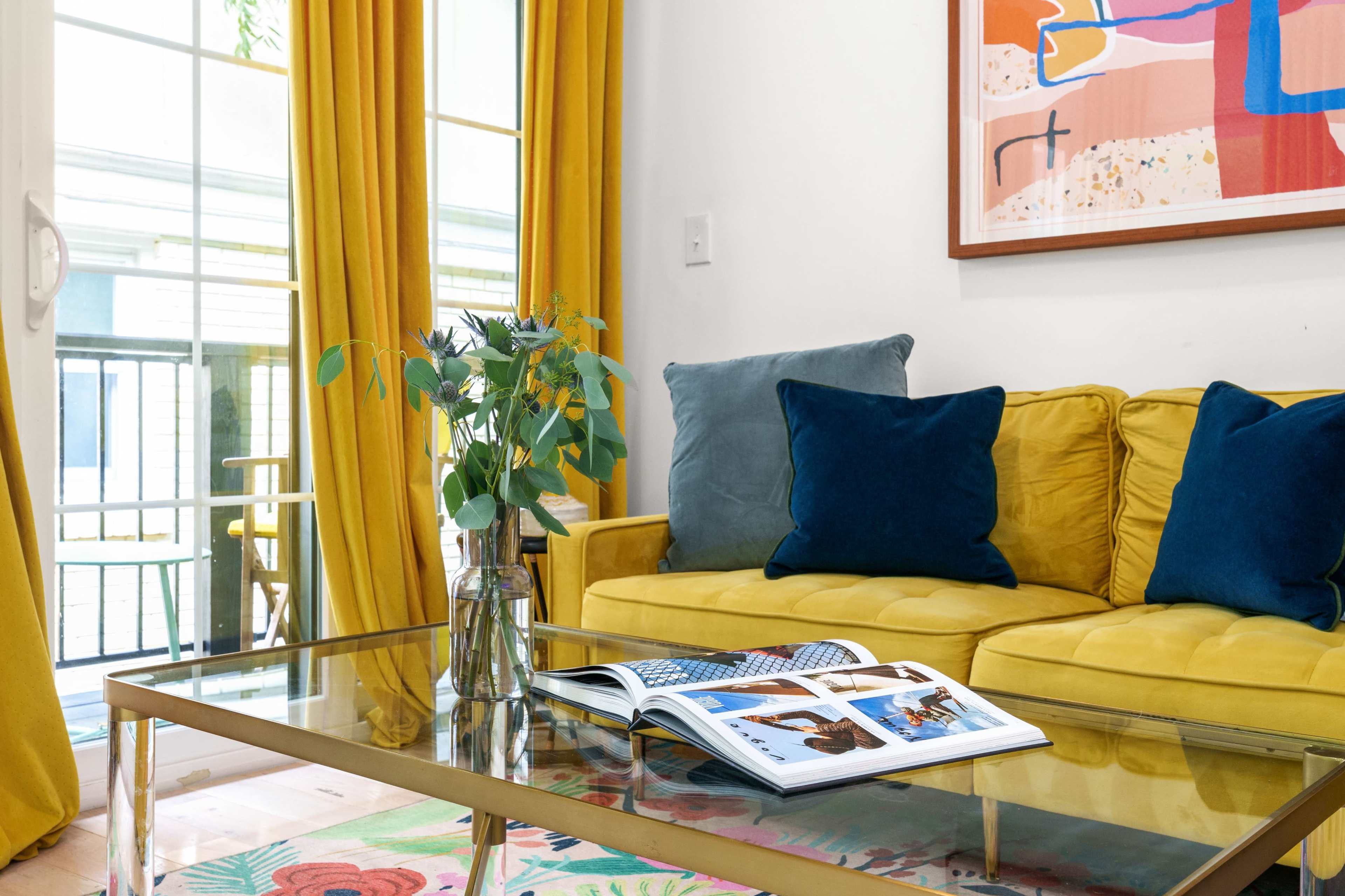 The scene features a bright living room with a yellow couch, blue pillows, a glass coffee table, and a decorative bouquet, alongside a colorful rug and a framed artwork on the wall.