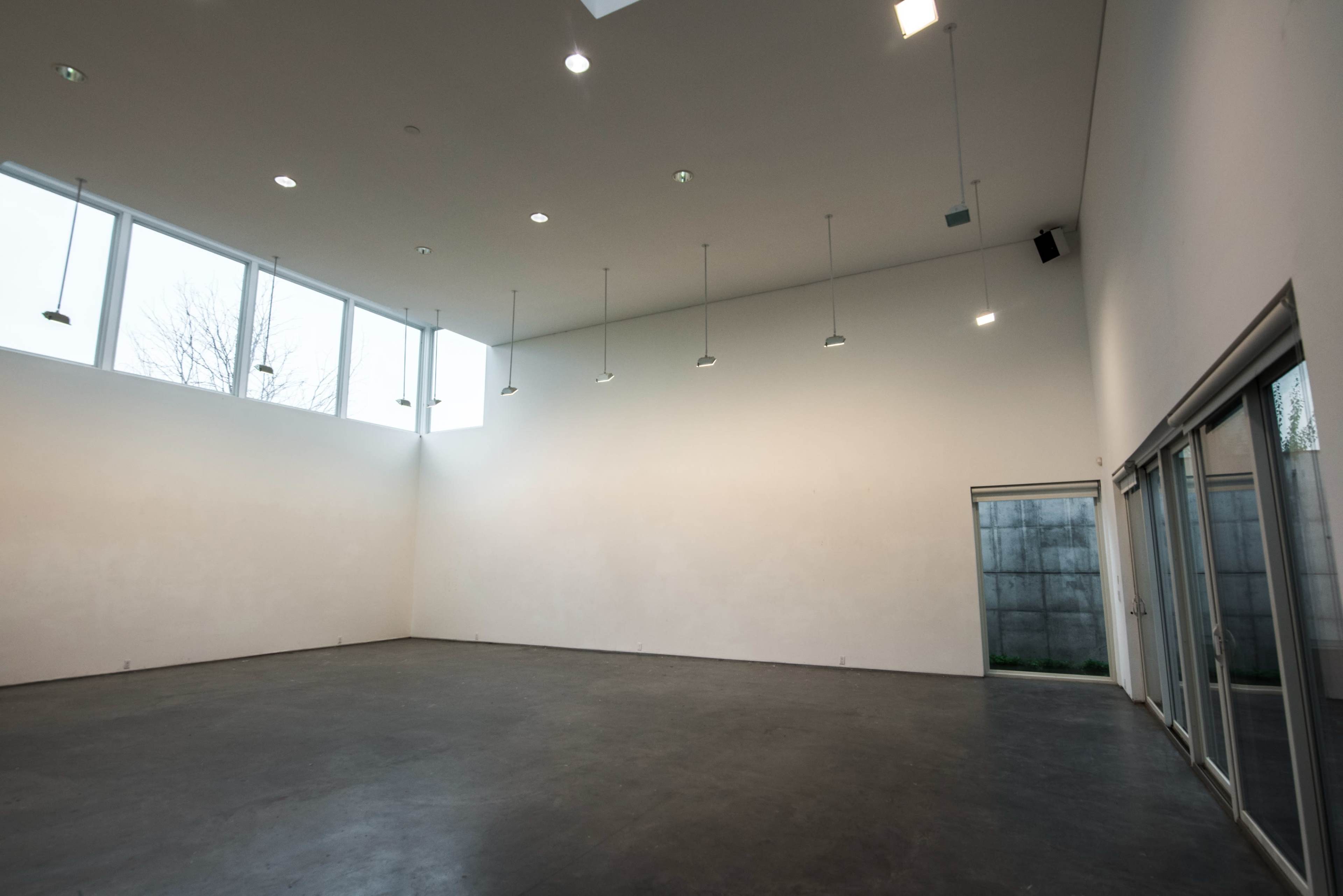 The image shows a spacious, empty room with high ceilings and large windows allowing natural light to enter.