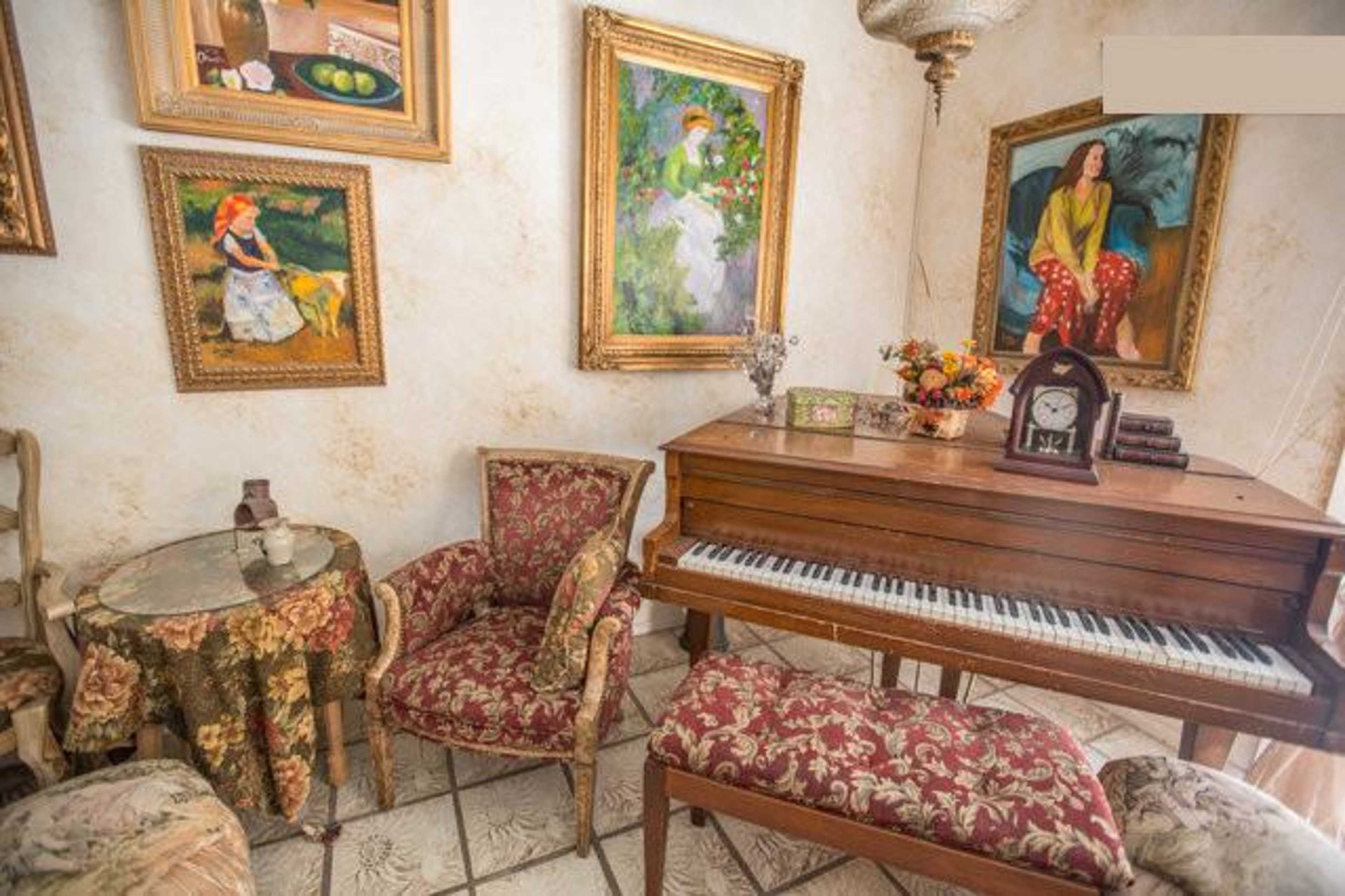A cozy room featuring a wooden piano, patterned chairs, a small round table, and several framed paintings on the walls.