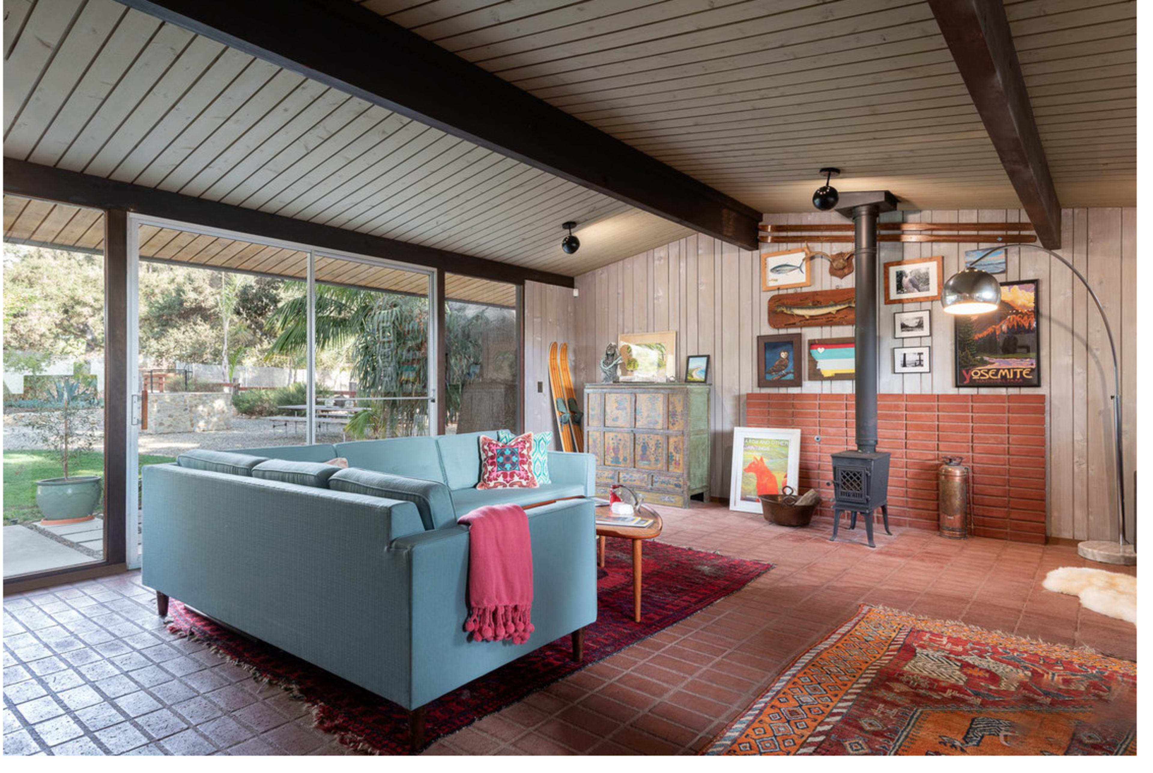 The image shows a cozy living room featuring a light blue sofa, a wood-burning stove, and various framed artworks on the walls, with large glass windows offering a view of a garden.