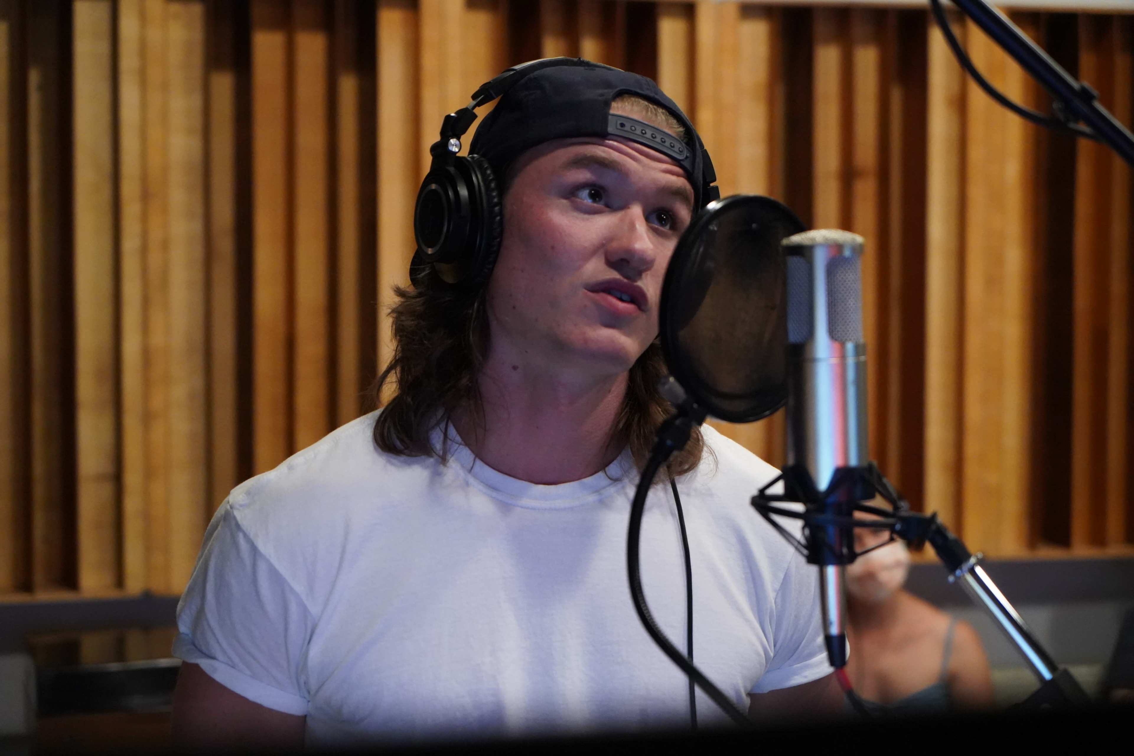A young man with long hair wearing a white t-shirt and headphones speaks into a microphone in a recording studio.