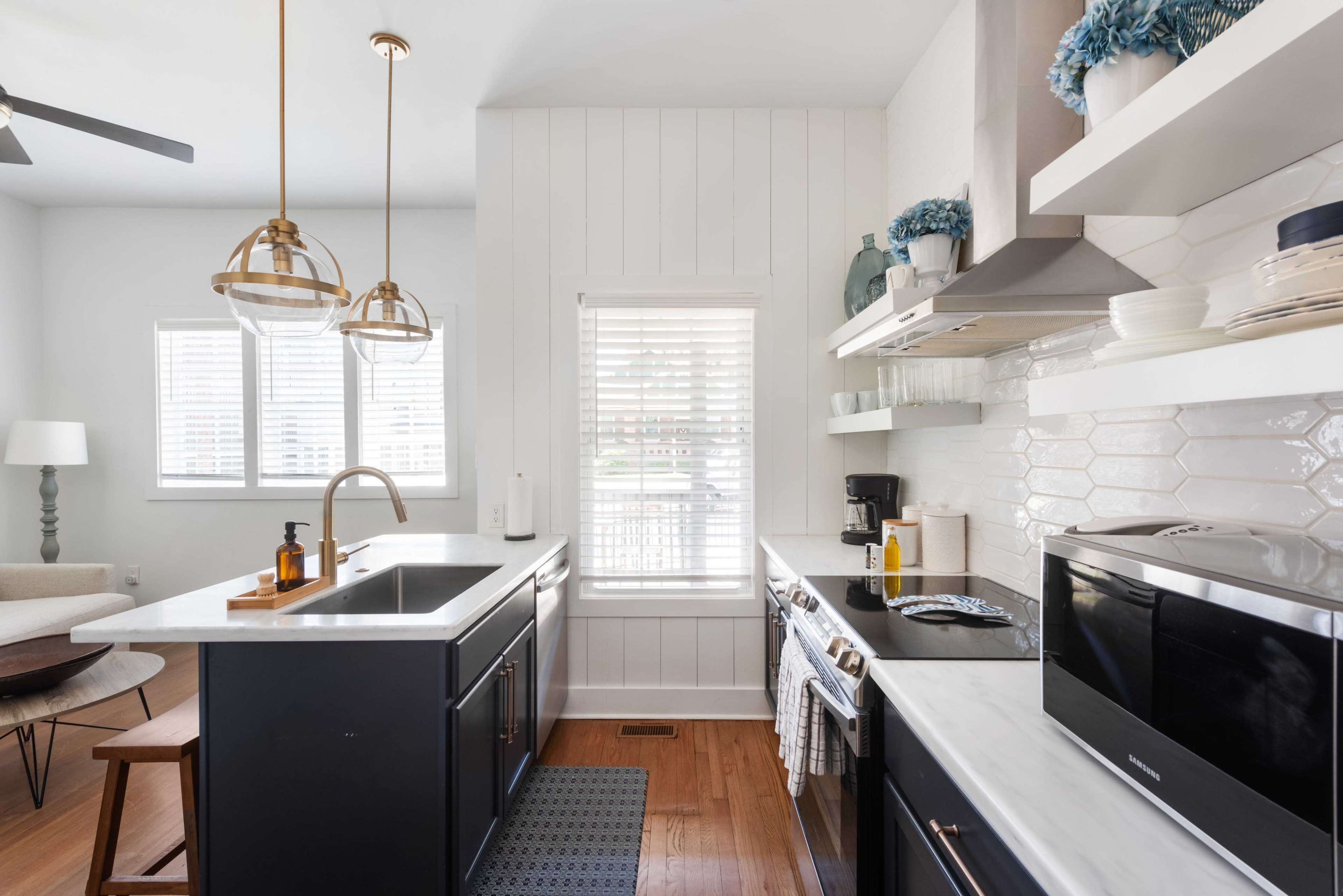 A modern kitchen features dark cabinetry, white walls, and a large window with blinds, alongside stainless steel appliances and decorative items on the shelves.