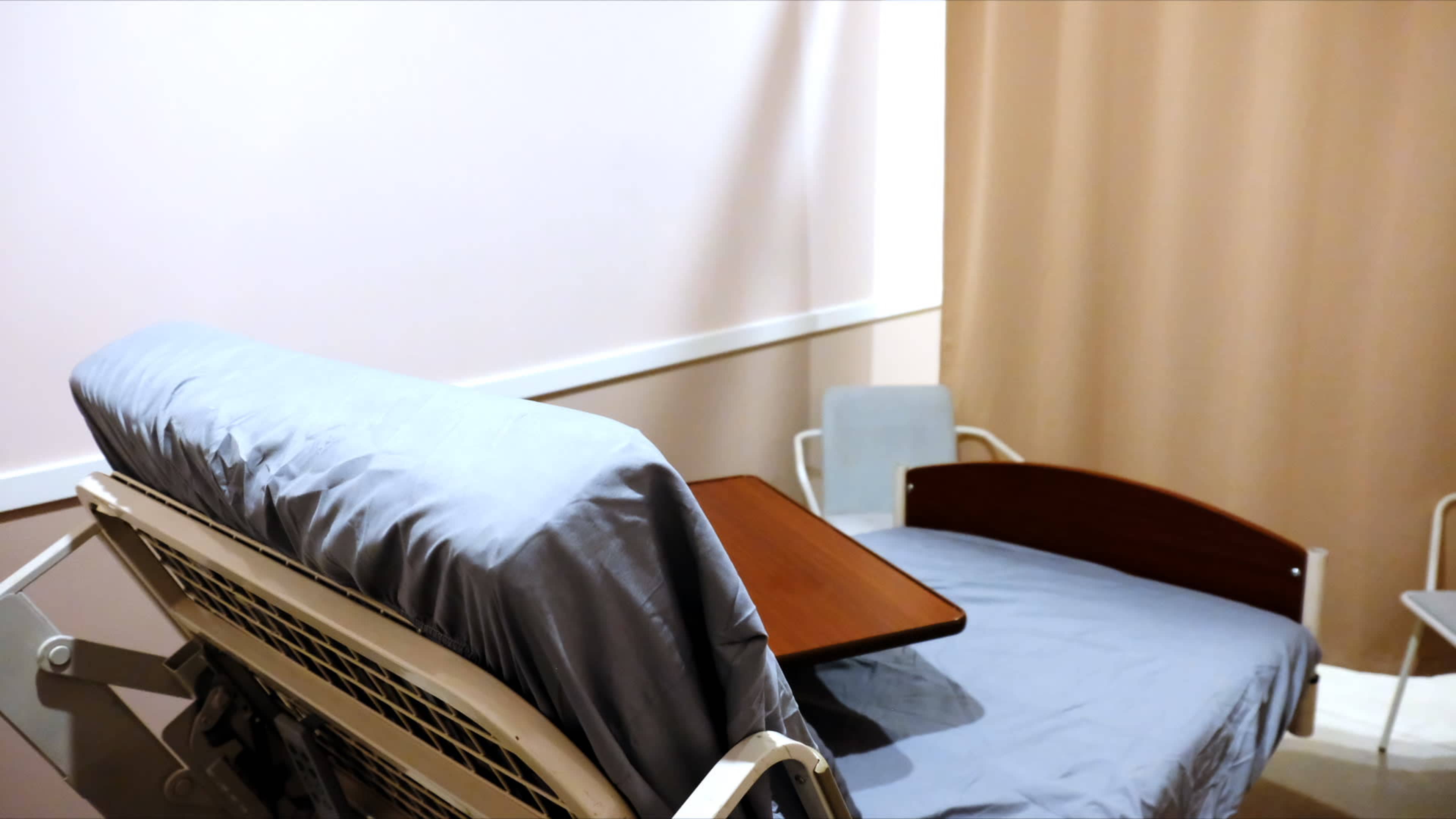 The image shows a hospital room with a bed featuring a light blue sheet and a wooden table attached to the side.