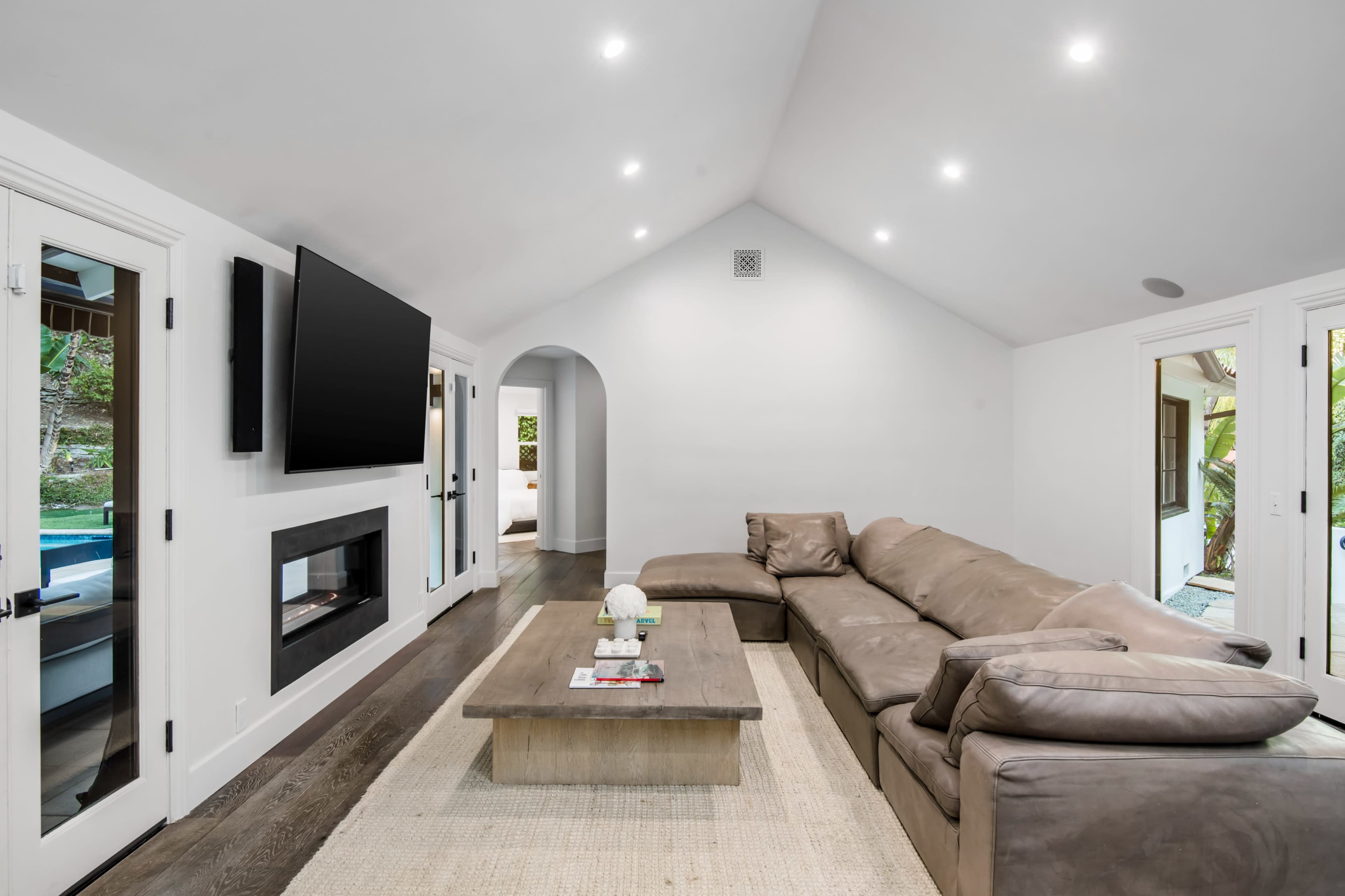 The spacious living room features a large sectional sofa, a coffee table, a flat-screen TV mounted on the wall, and a clean, modern design with natural light flowing in.