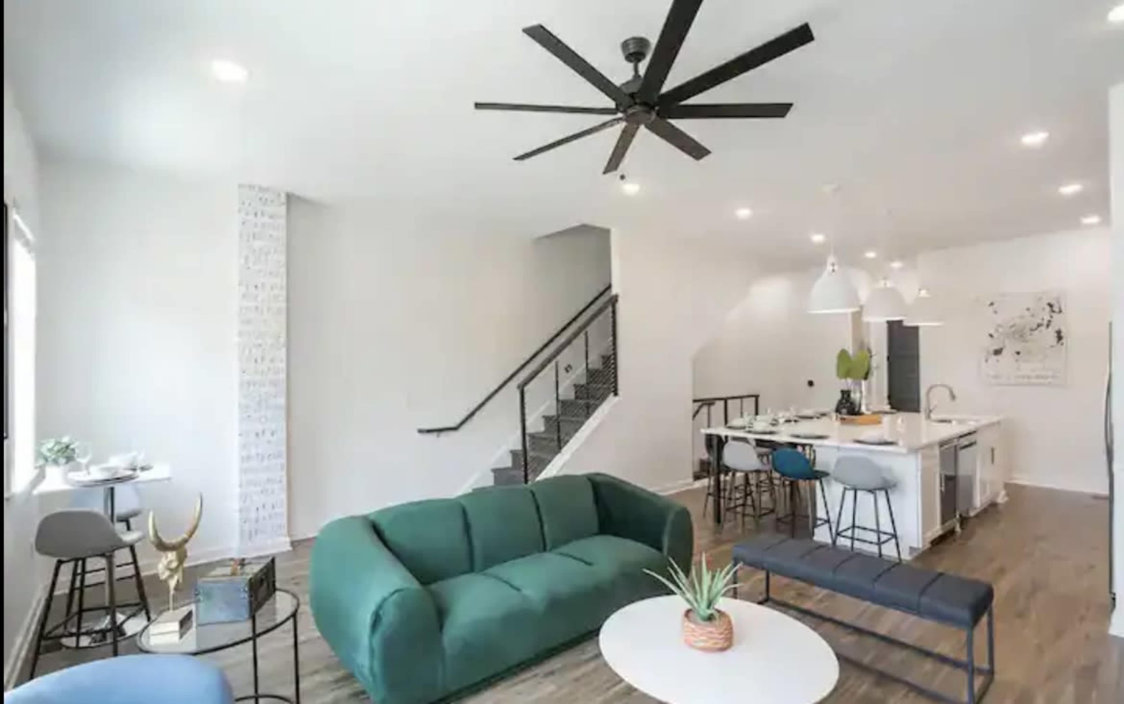 A modern living space with a green sofa, a round coffee table, a kitchen area featuring bar stools, and a staircase in the background.