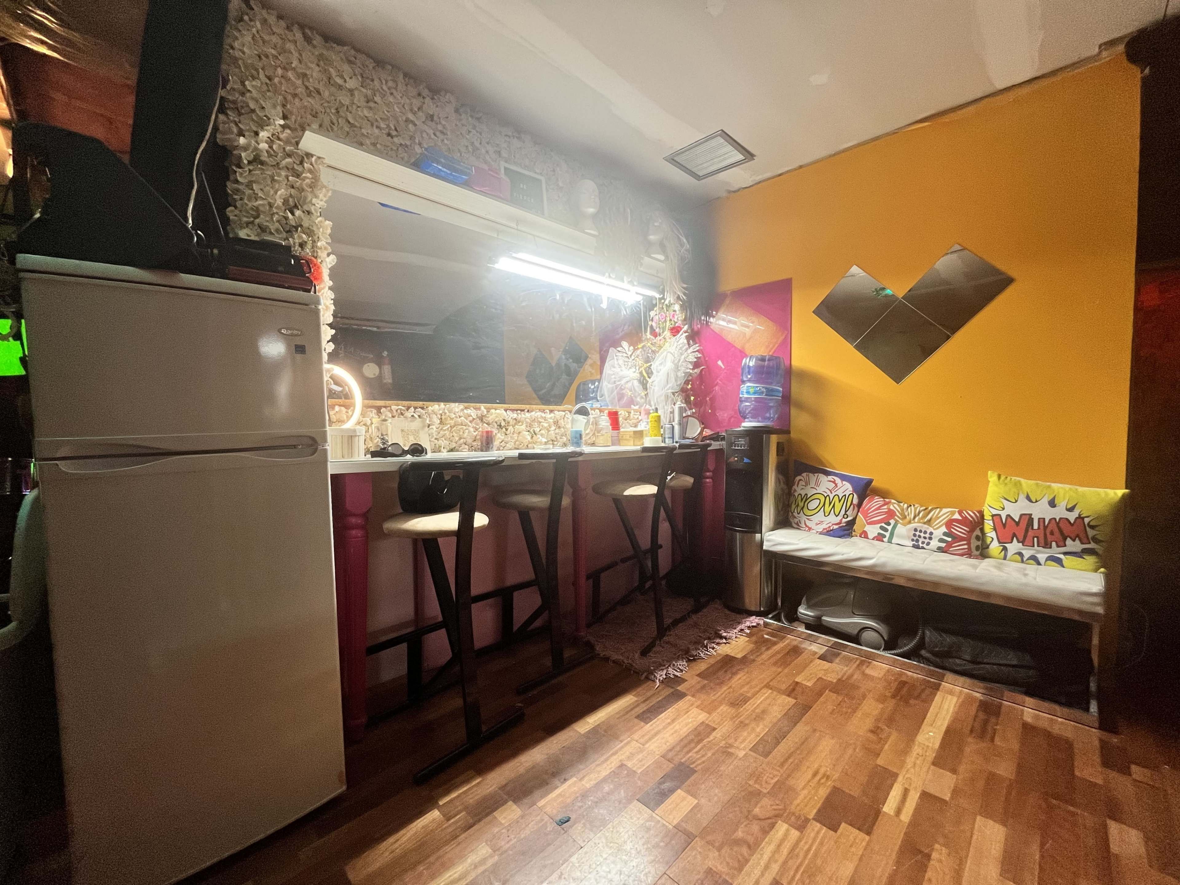 A small kitchen area with a bar counter, stools, refrigerator, and a colorful wall featuring decorative cushions.
