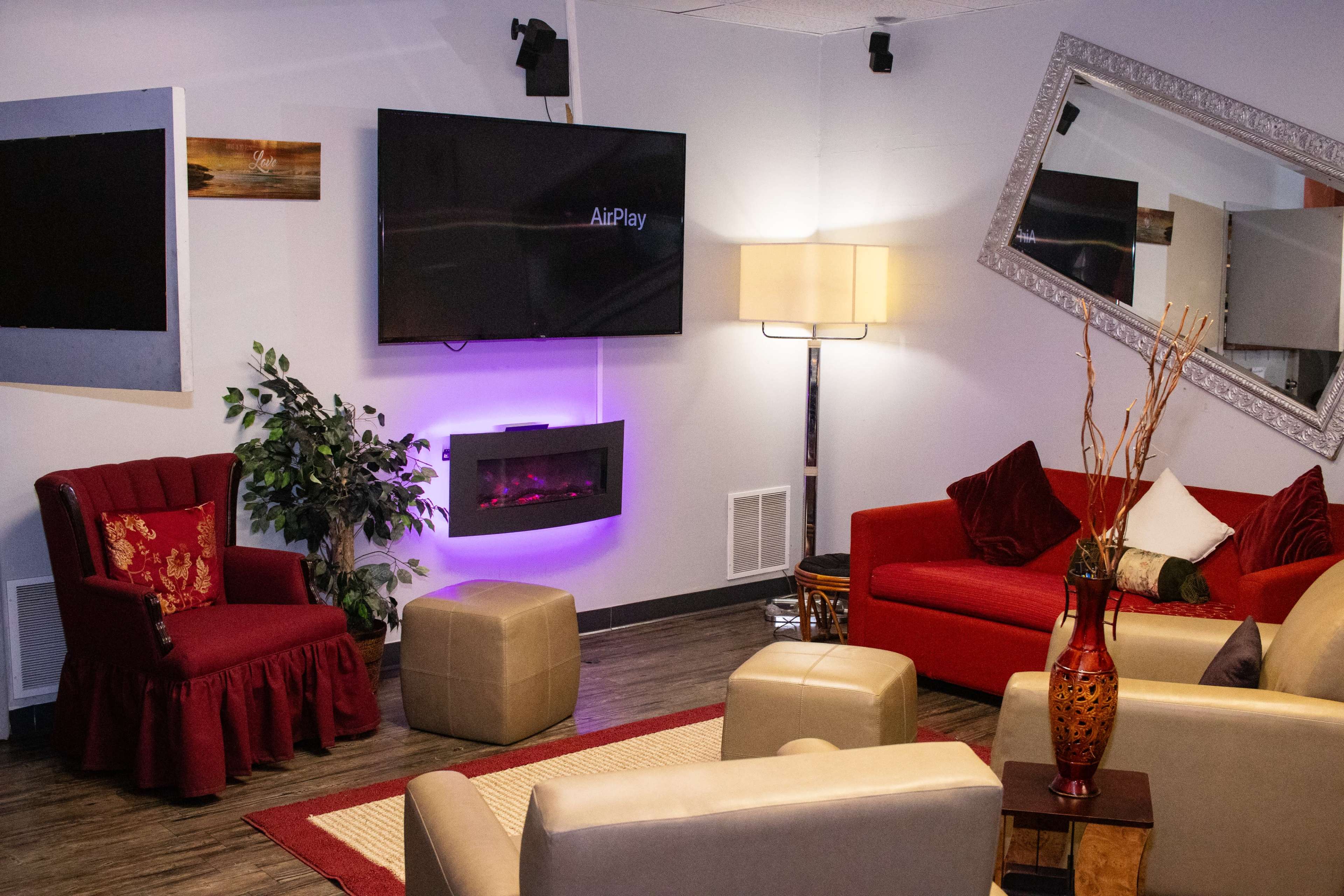 A cozy room features a TV mounted on the wall, a modern electric fireplace, a lamp, and various seating options including red and cream-colored chairs and ottomans.