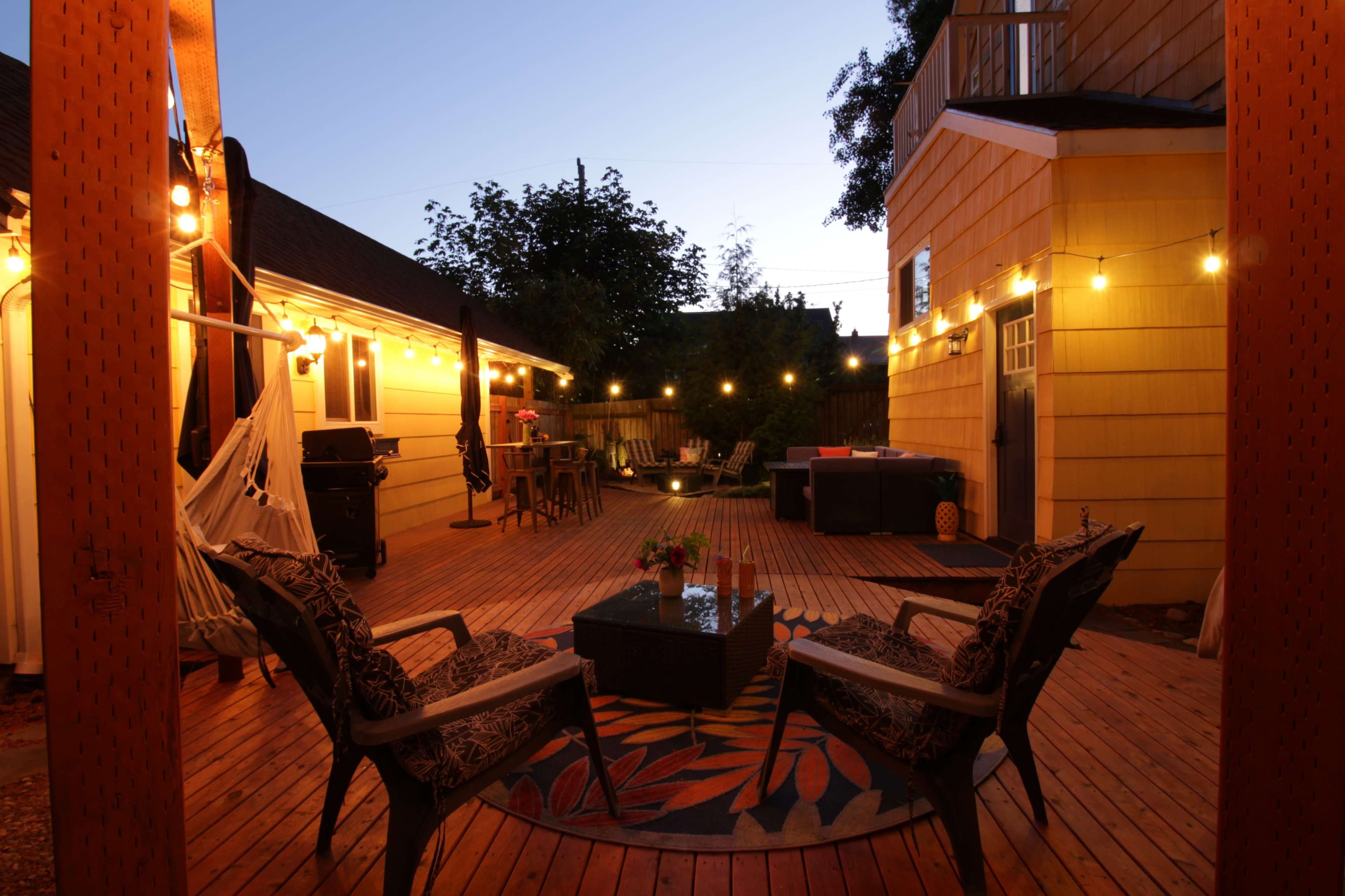 A wooden deck features two chairs and a table, illuminated by string lights, with a backdrop of two houses and greenery in the evening light.