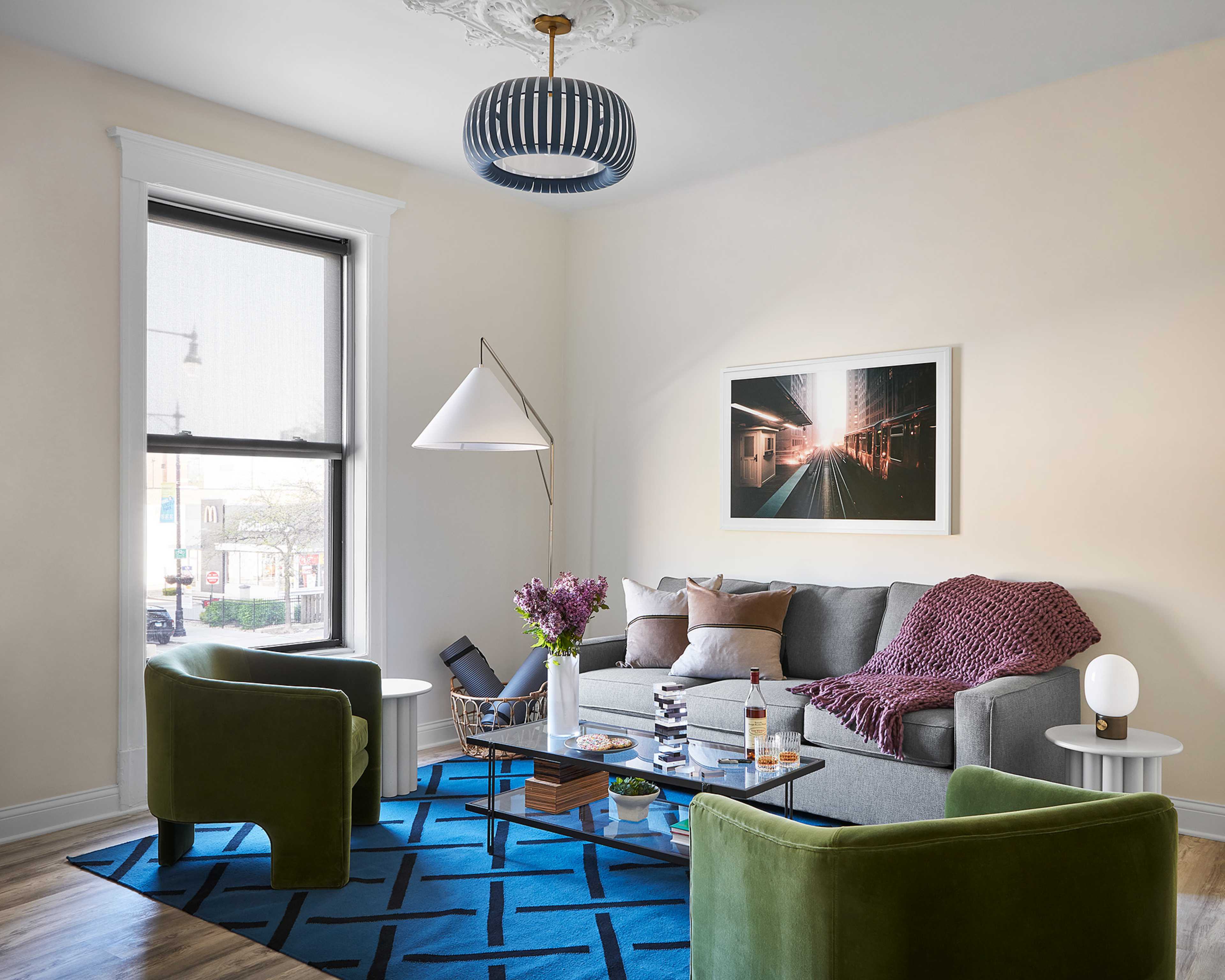 A modern living room features a gray sofa with patterned pillows, two green armchairs, a coffee table with decorative items, and a patterned rug under a ceiling light and artwork.