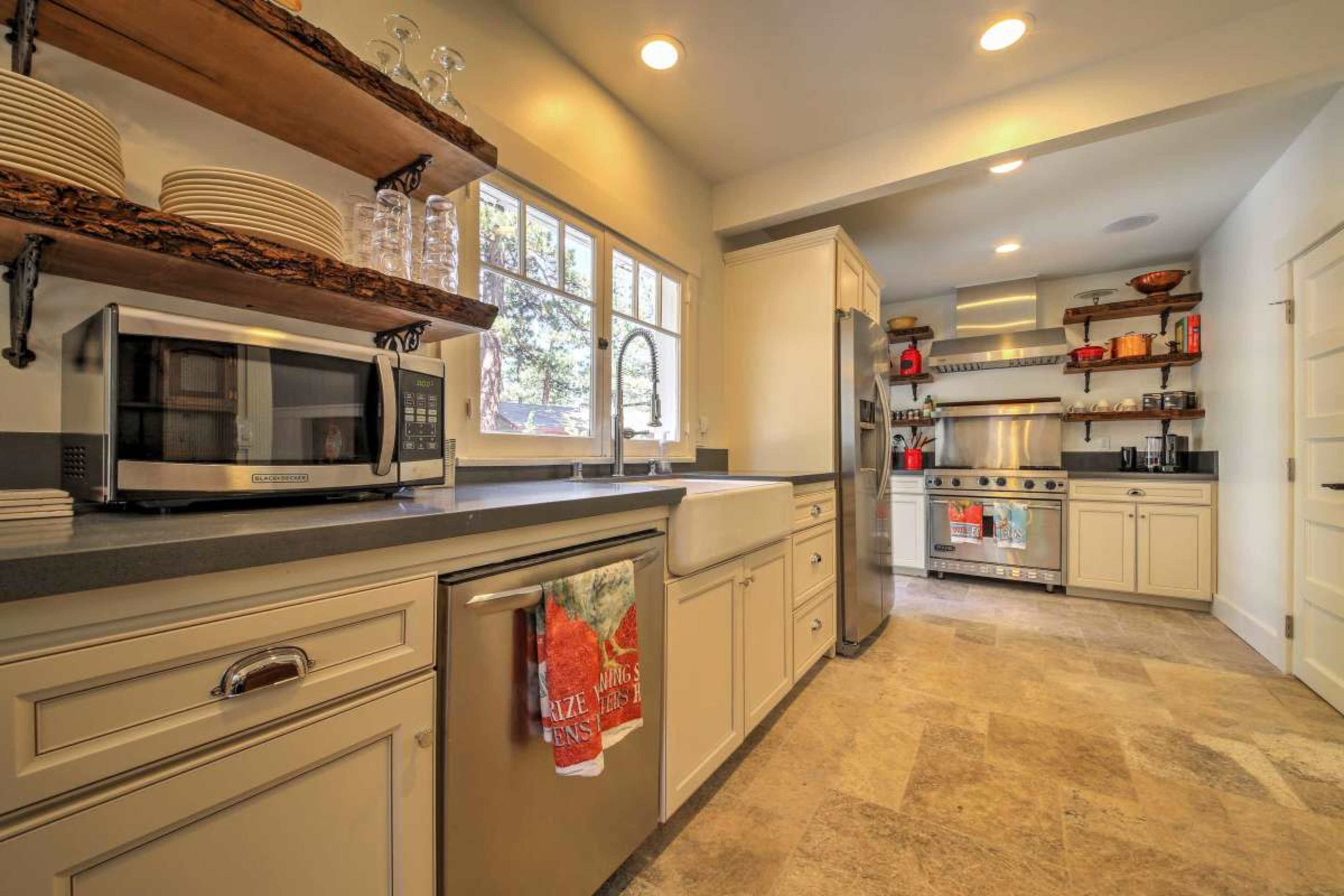 A modern kitchen featuring stainless steel appliances, a farmhouse sink, open shelving, and a combination of light cabinetry and stone flooring.