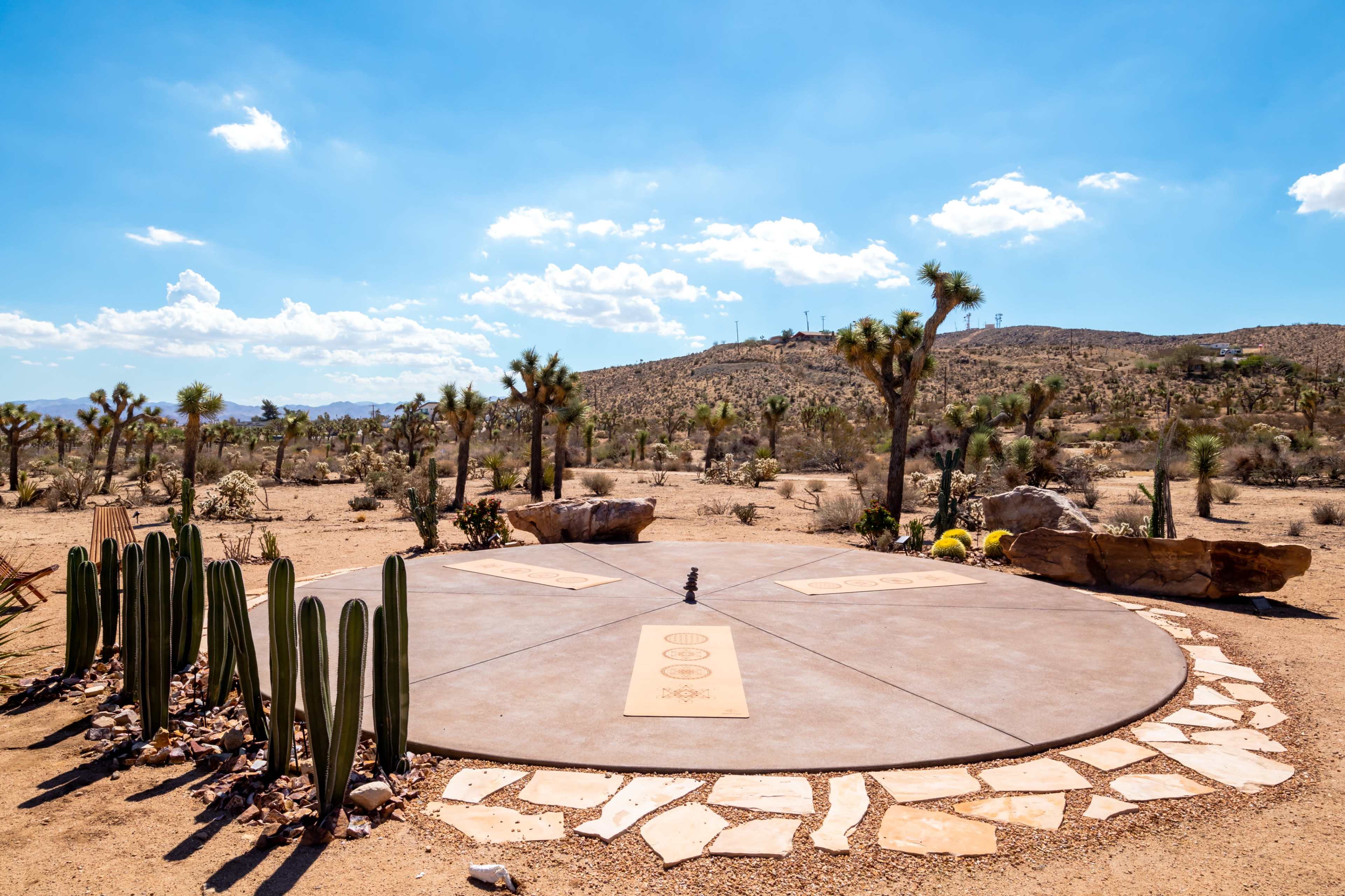 A circular stone platform is surrounded by cacti and desert vegetation, with a backdrop of hills and a blue sky.
