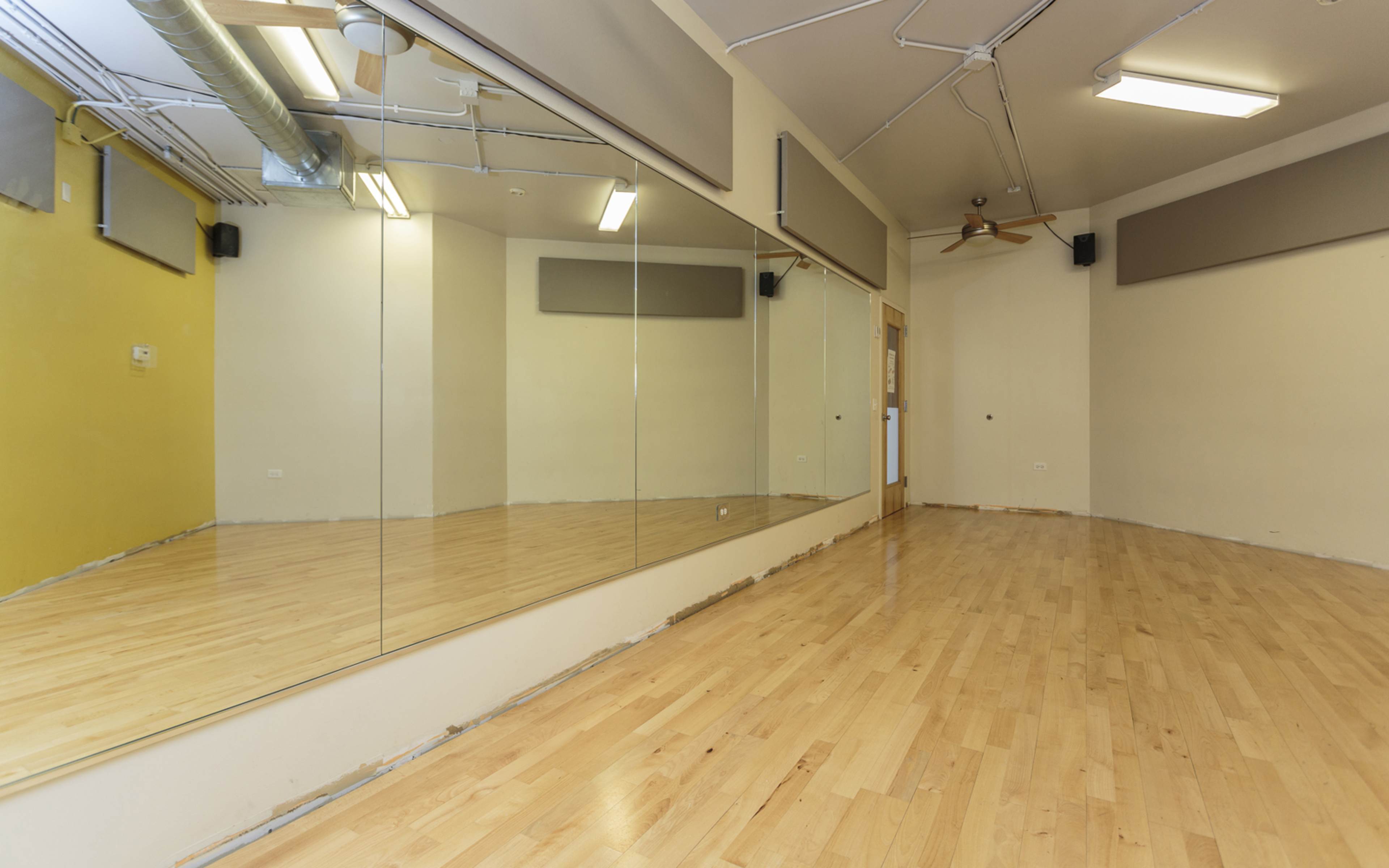 The image shows an empty room with wooden flooring and large mirrors on one wall.