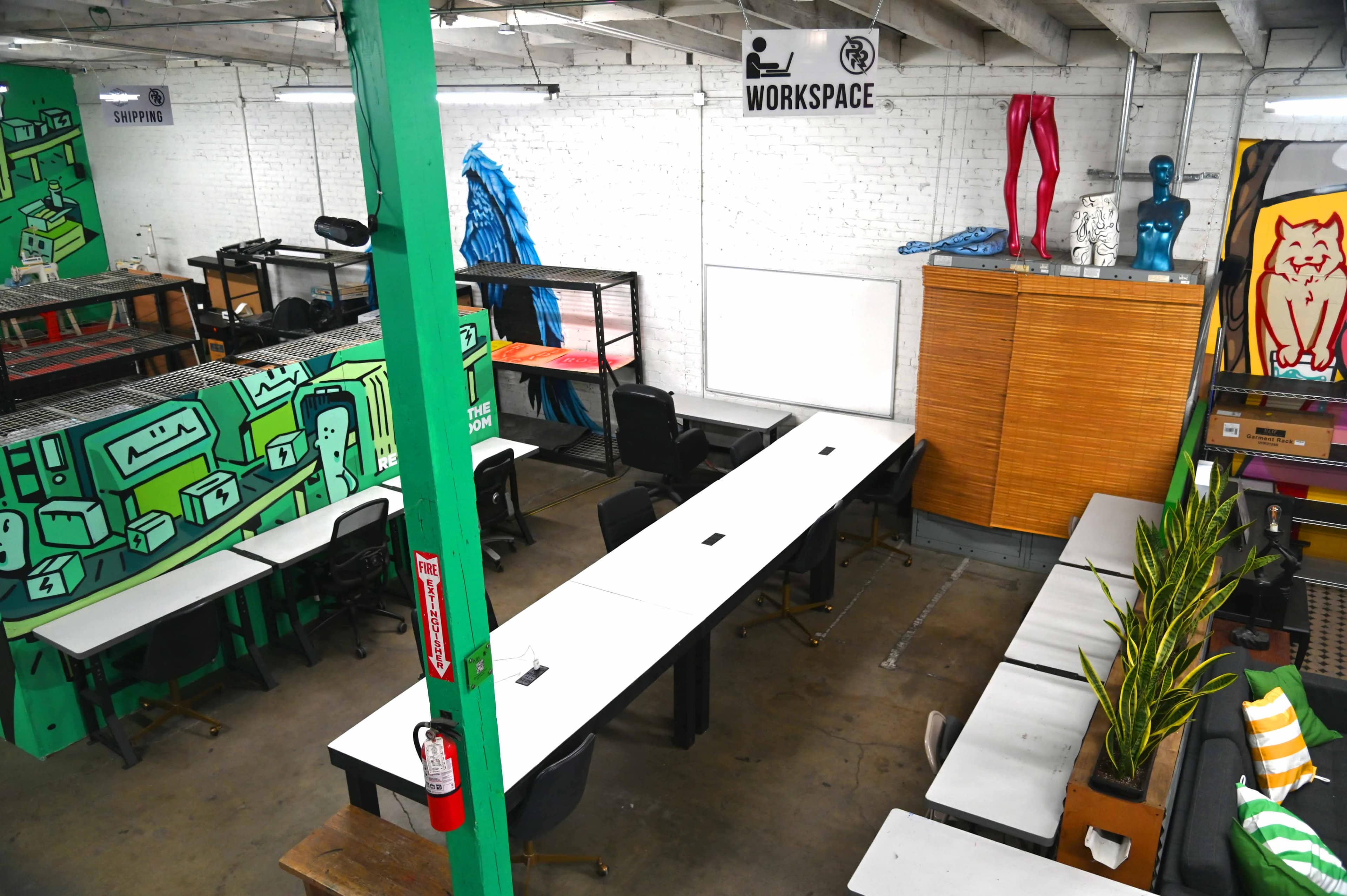 The image shows an open workspace with several long tables, chairs, and various colorful murals on the walls.