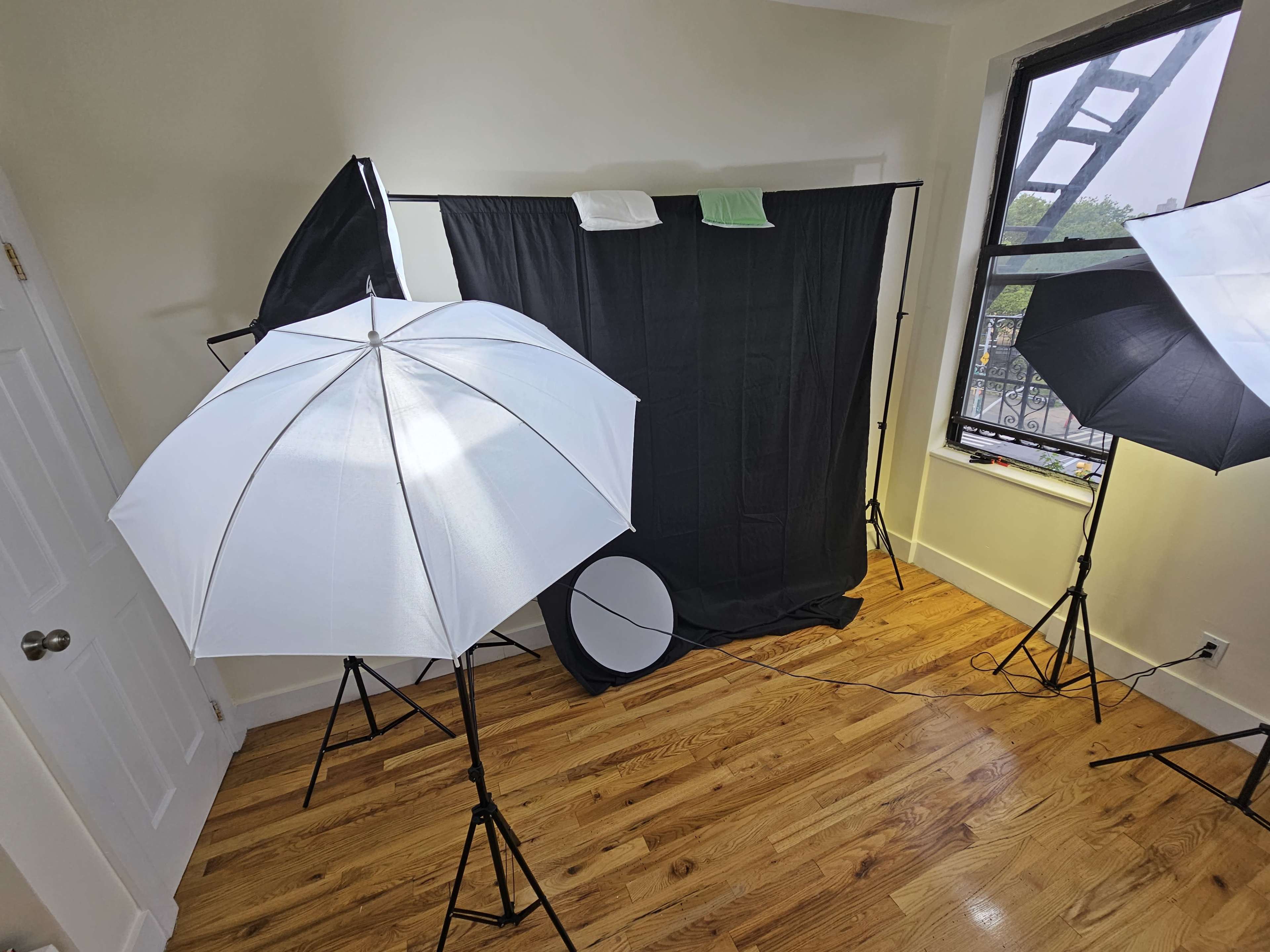 A photography setup features a black backdrop, multiple lighting umbrellas, and a round reflector in a room with wooden flooring and a window.