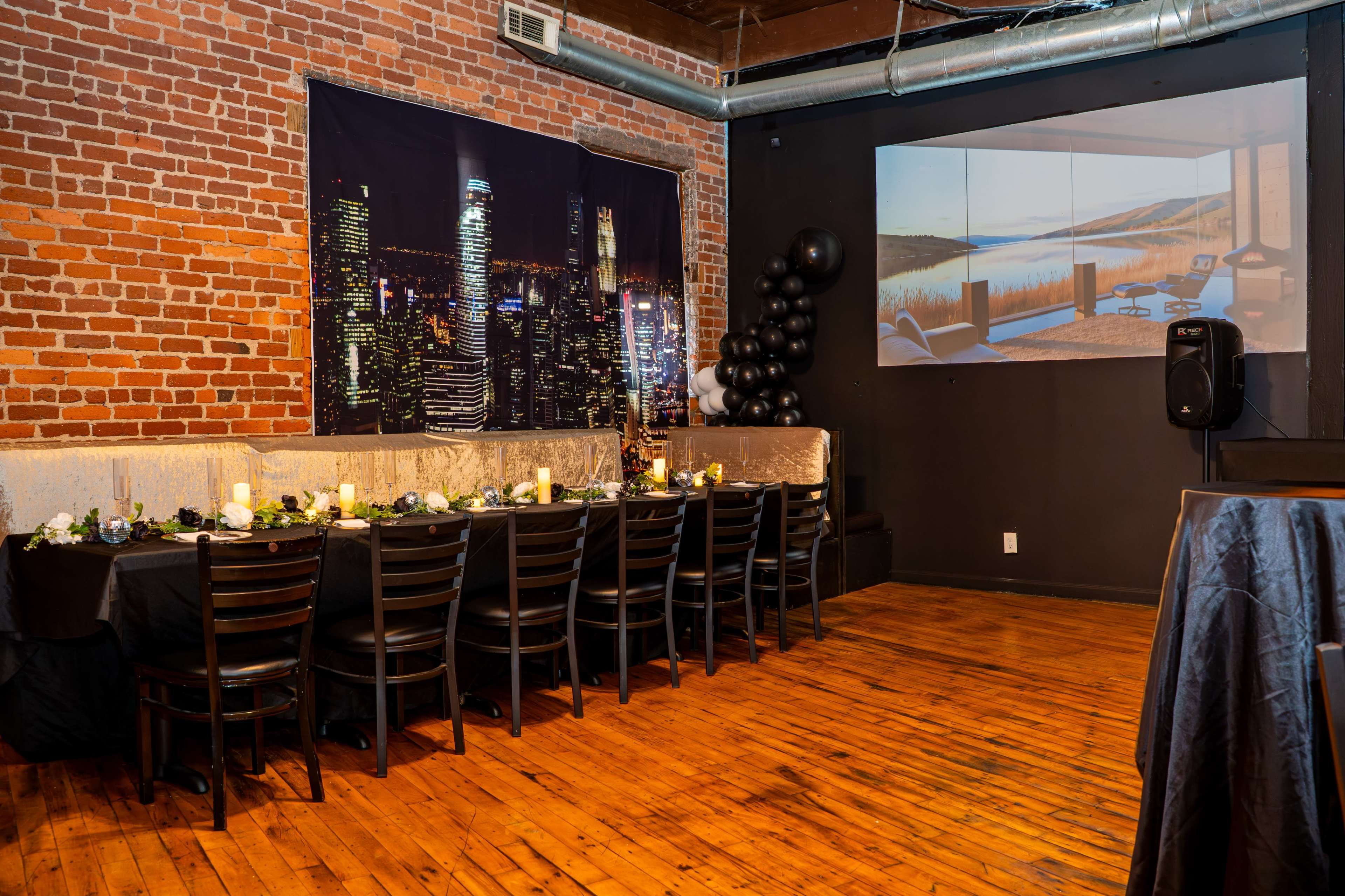 The scene features a long table set for dining, accompanied by a brick wall and a video projection of a landscape on an adjacent wall.