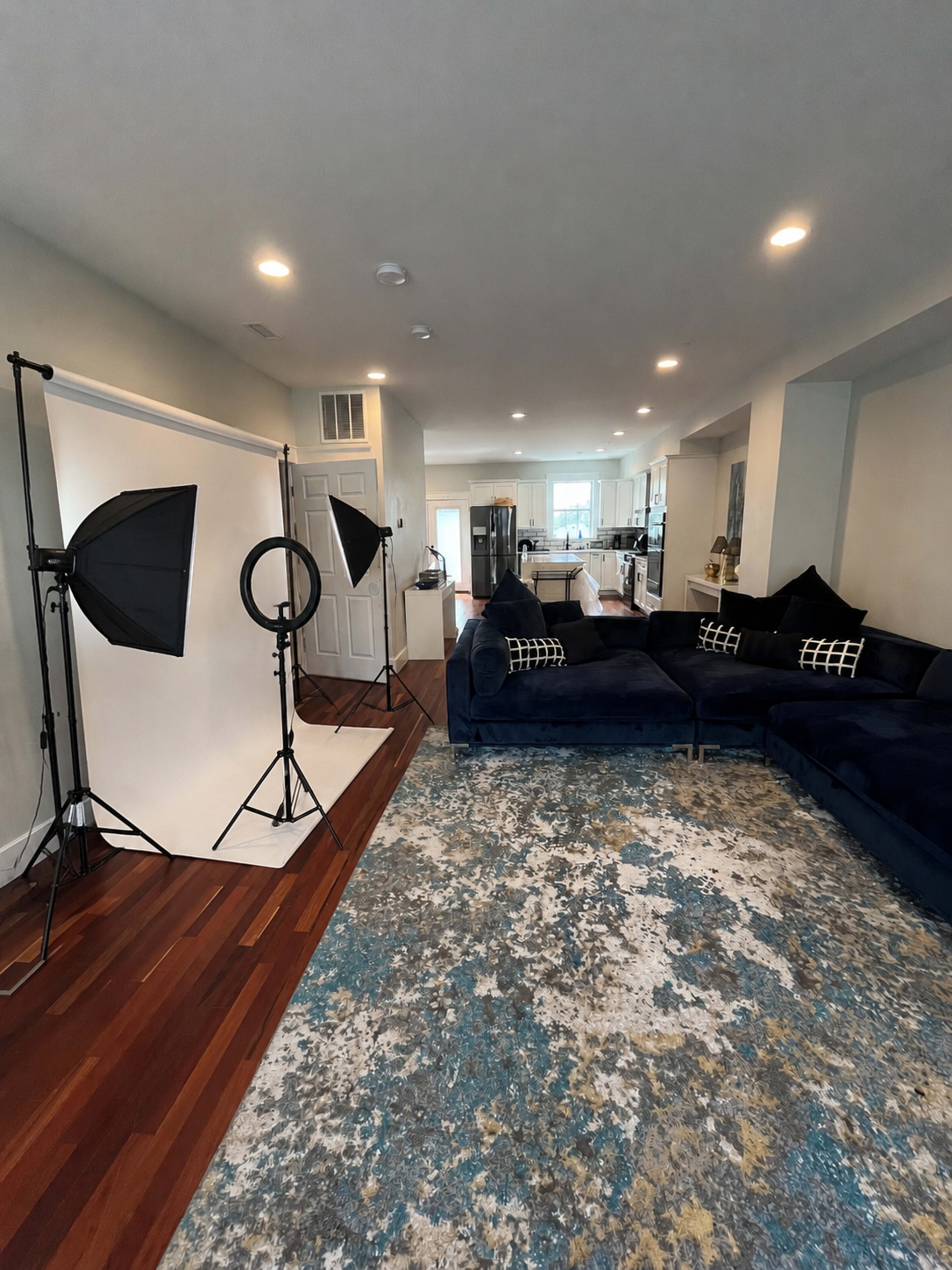The image shows a spacious living area with a blue sectional couch, a light rug, and photography equipment set up on one side.