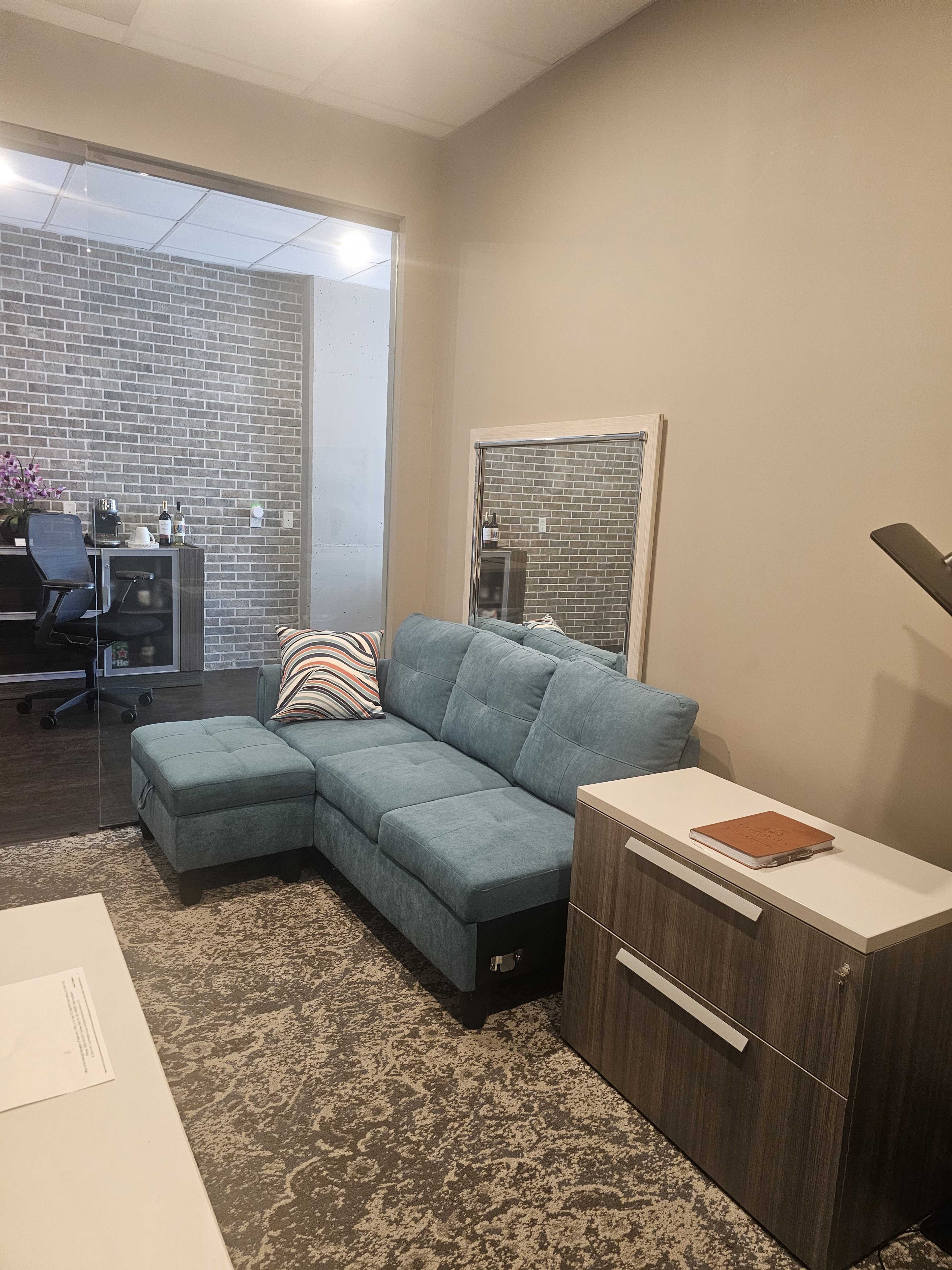 The image shows a modern office space featuring a teal sectional sofa, a mirror, and a white cabinet beside it, with a brick wall visible in the background.