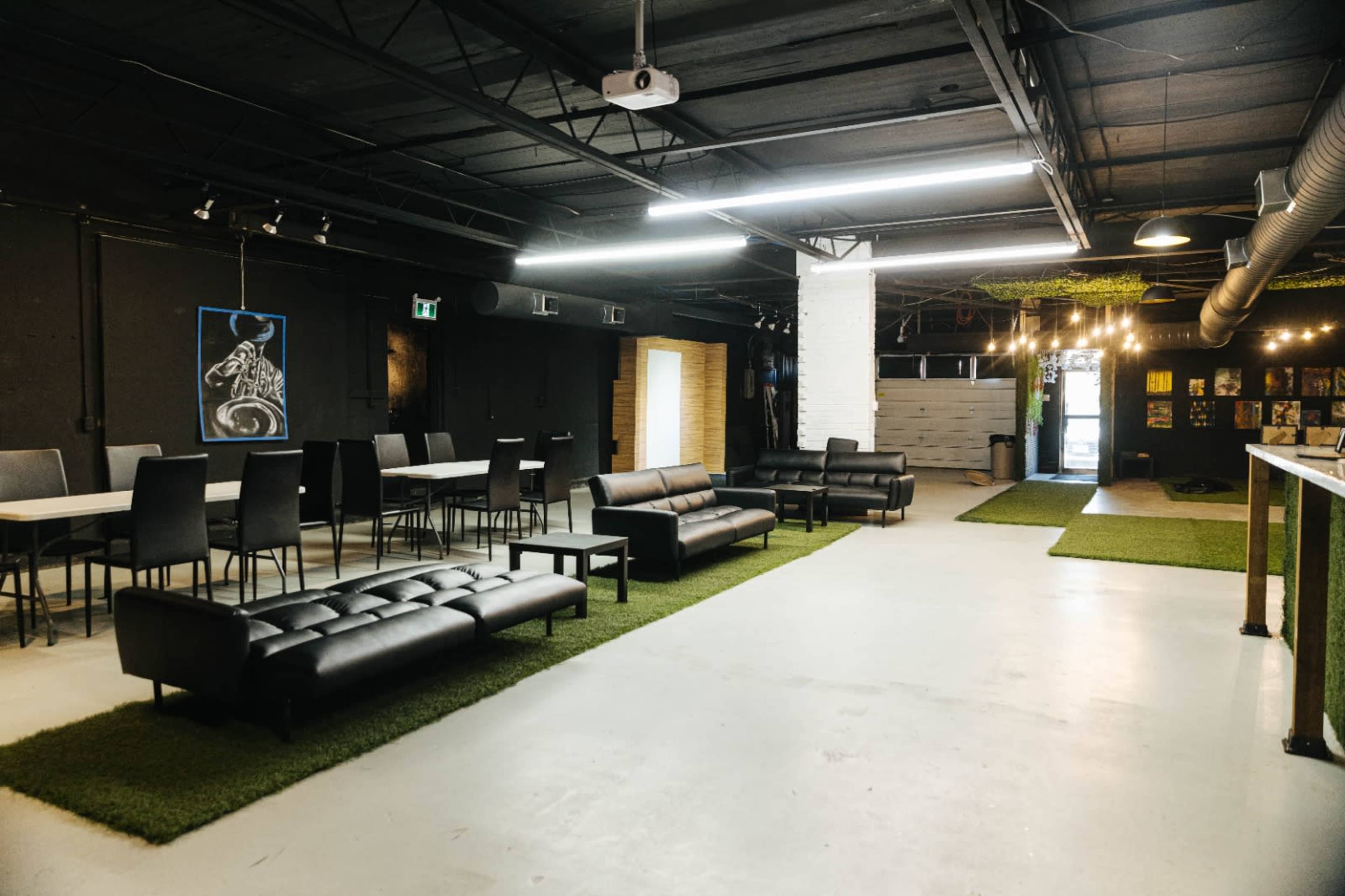 The image shows a spacious interior with black walls, a few tables set with chairs, leather couches, and sections of artificial grass on the floor.