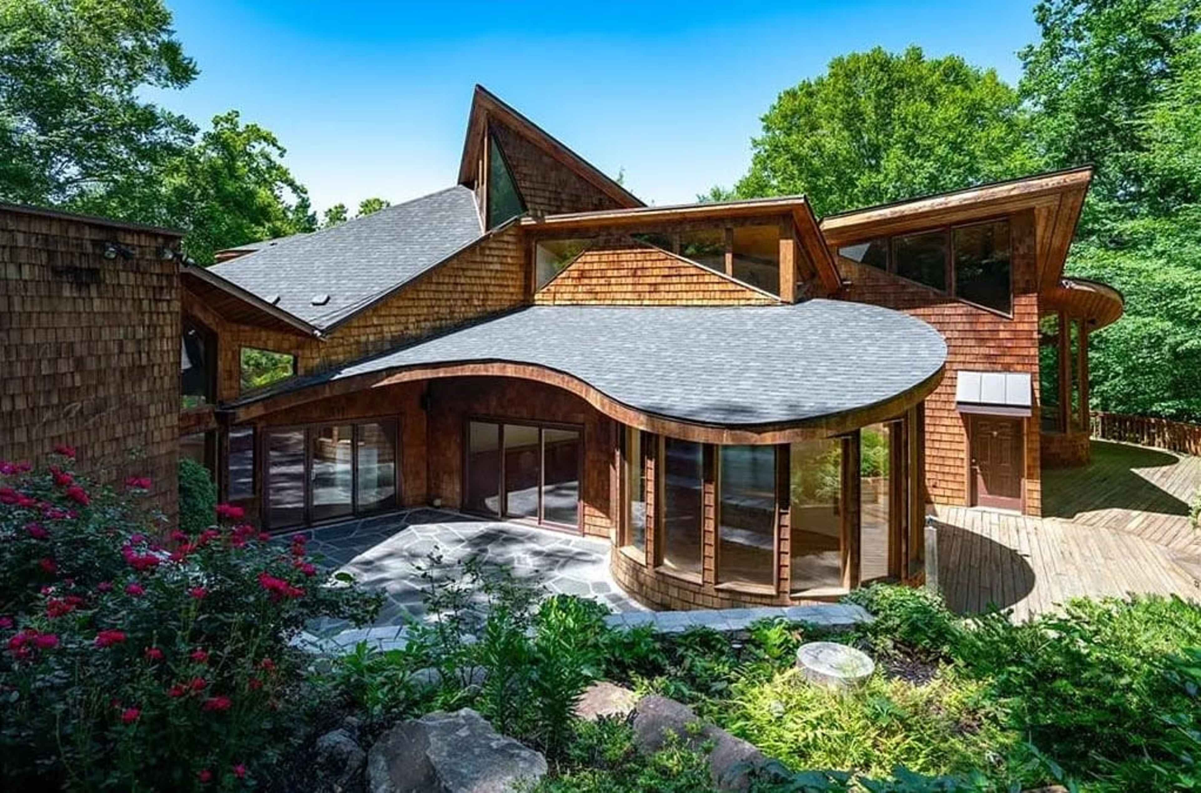 The image shows a modern wooden house with a curved design, large glass windows, and a surrounding garden with trees and flowers.