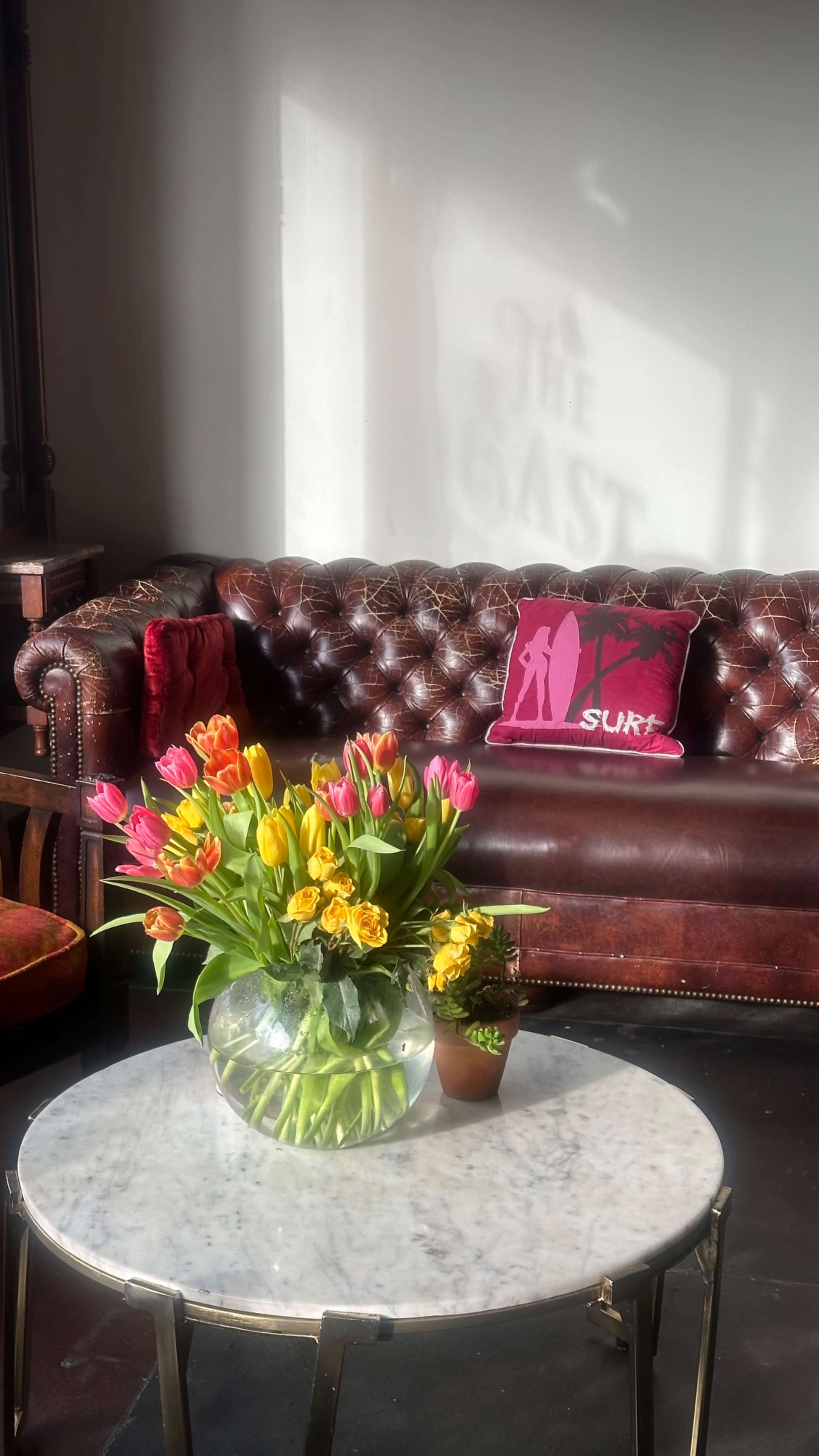 A brown leather tufted couch is positioned next to a marble table adorned with a large bouquet of colorful flowers and a small potted plant, while a shadow on the wall displays the word "CAST."