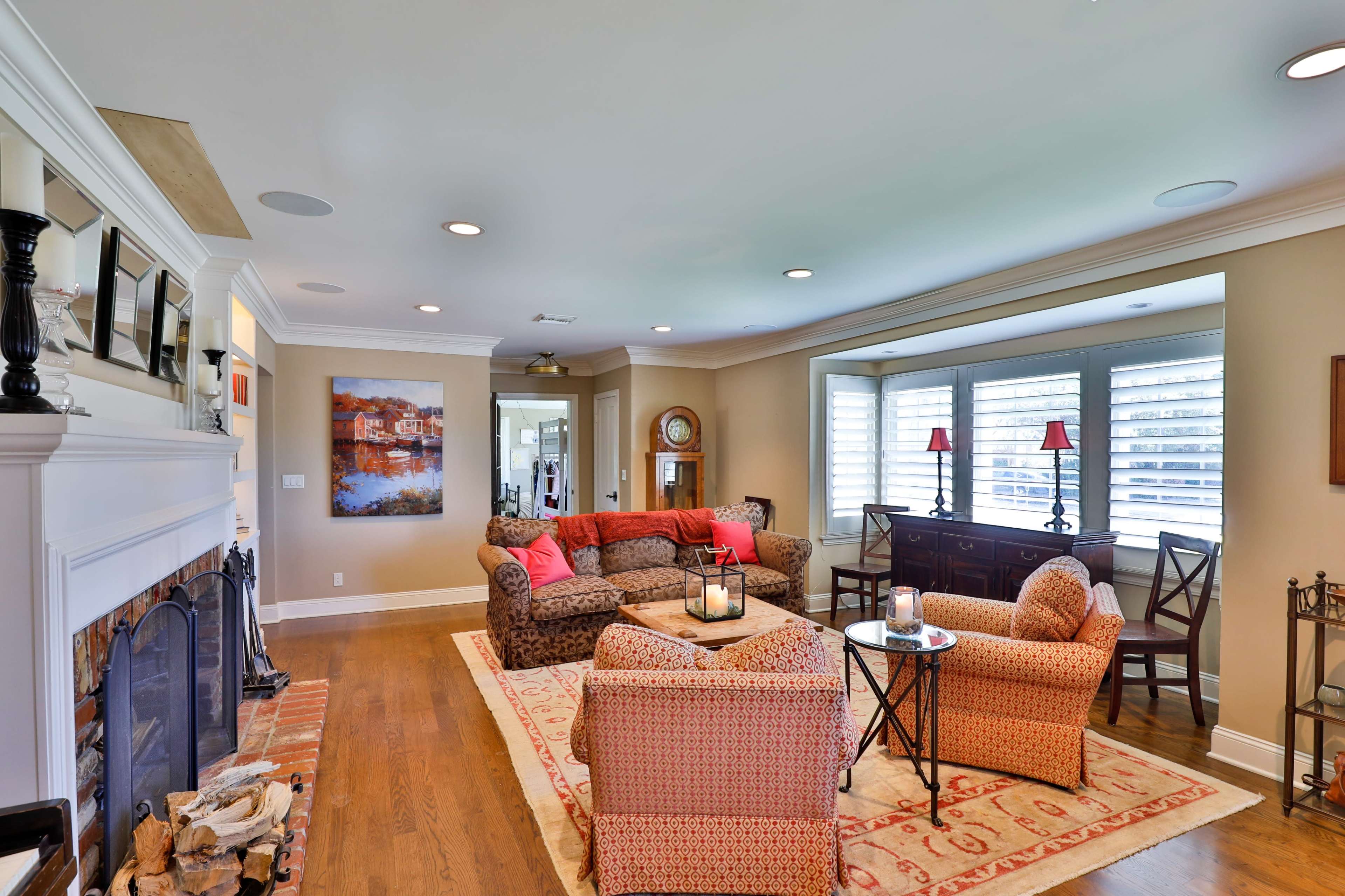 A cozy living room with a seating area featuring a patterned sofa and armchairs, a fireplace, and natural light coming through window shutters.