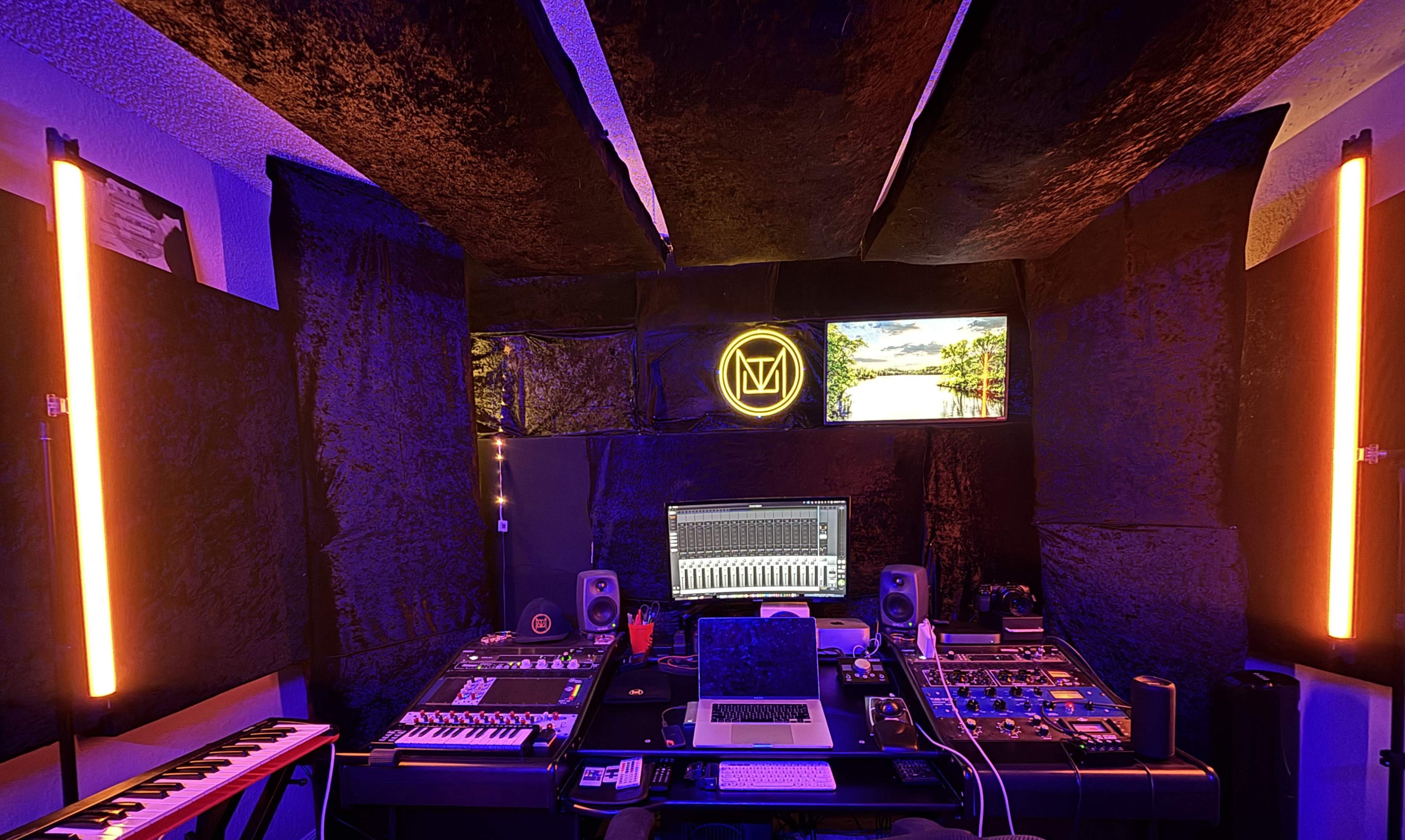 The image shows a modern music studio featuring a computer setup, audio equipment, and neon lighting in a dark, soundproofed room.