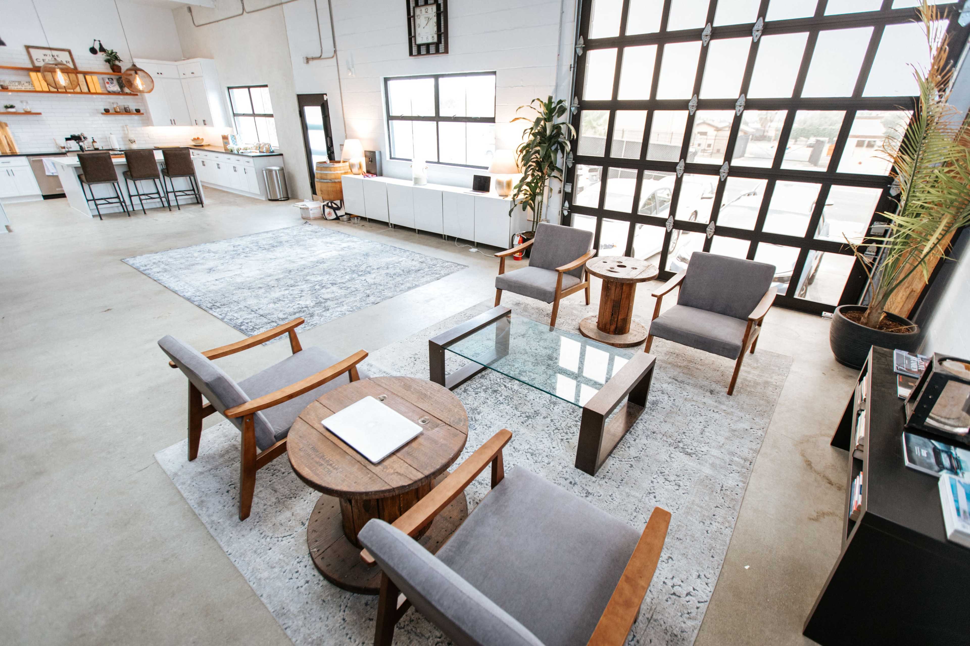 A spacious, modern living area with a kitchen in the background, featuring a seating arrangement of gray chairs and wooden tables on a light-colored rug.