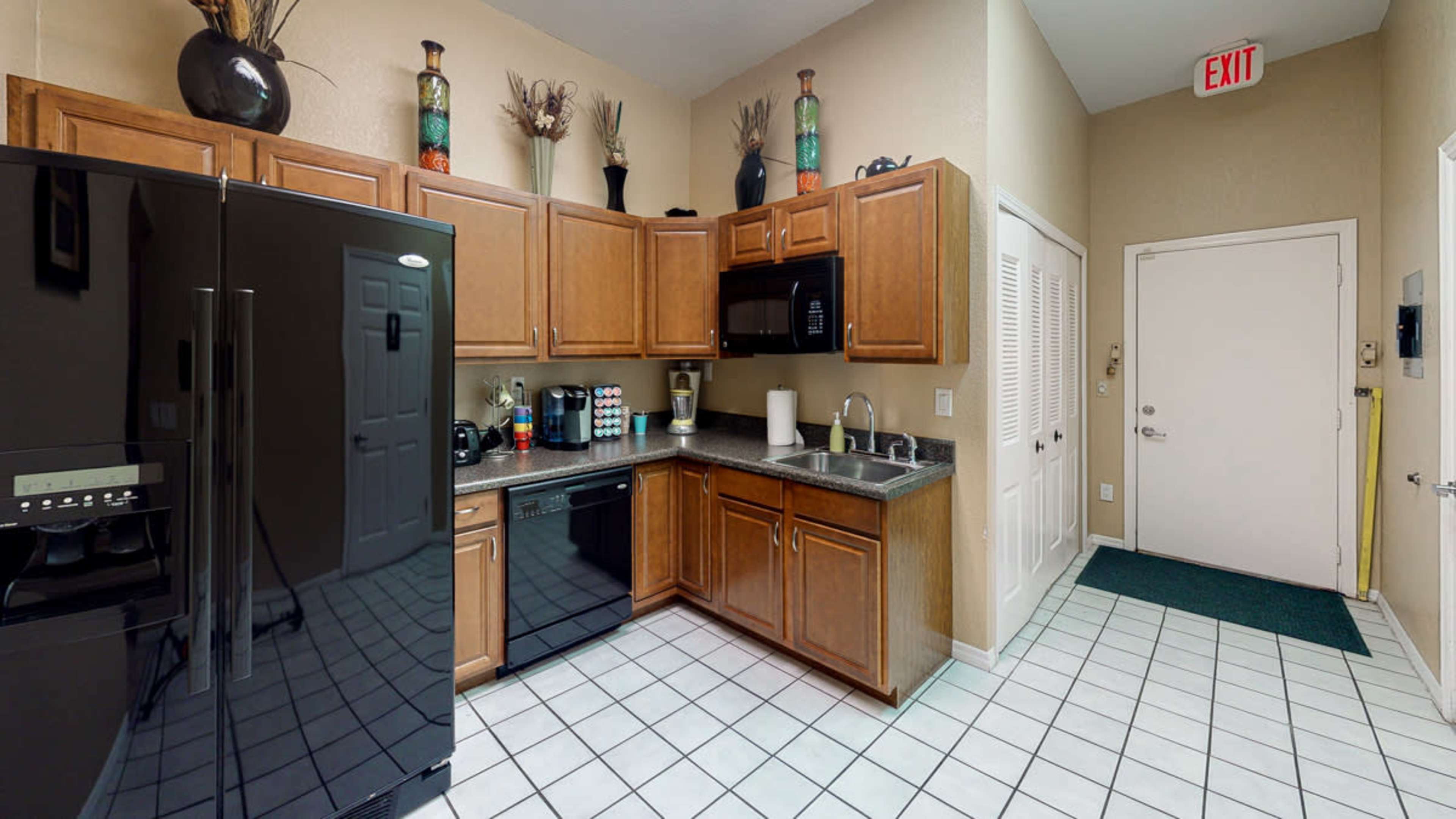 The image shows a kitchen equipped with wooden cabinets, a black refrigerator, a microwave, and a tiled floor, adjacent to a door leading outside.