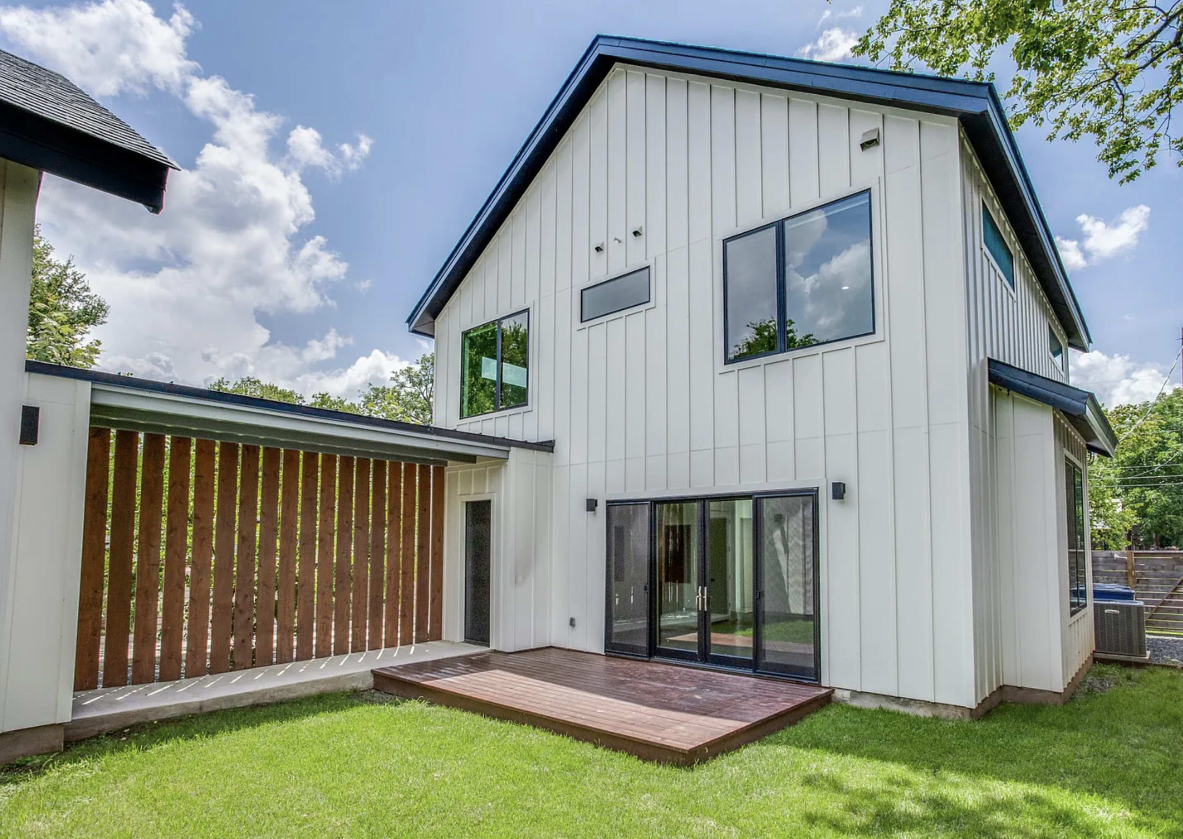 A modern two-story house features white vertical siding, large windows, and a wooden deck in the backyard.