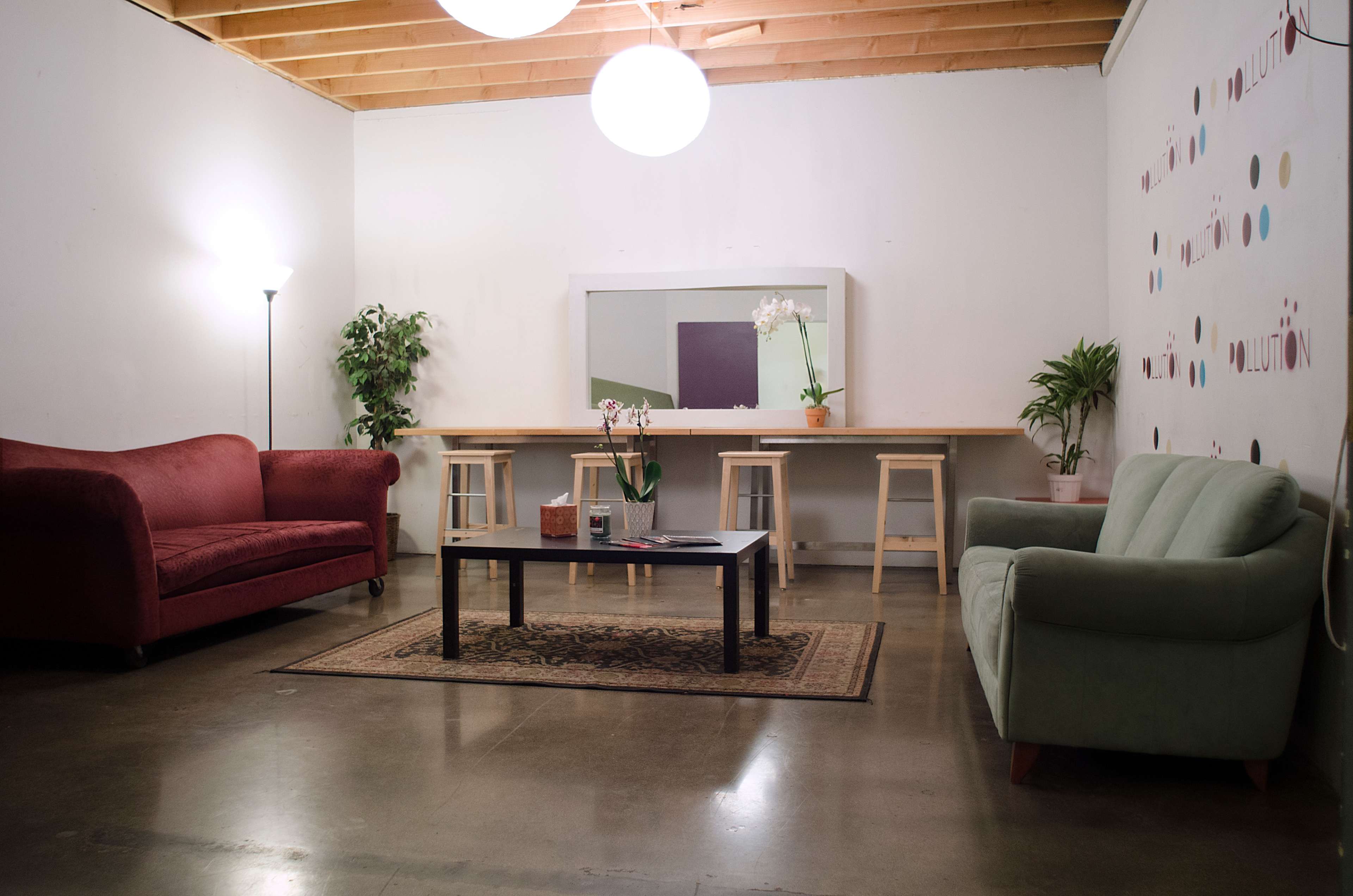 A minimalistic room features a red couch, a green couch, a rectangular coffee table, a long wooden table with stools, and a wall adorned with decorative dots.