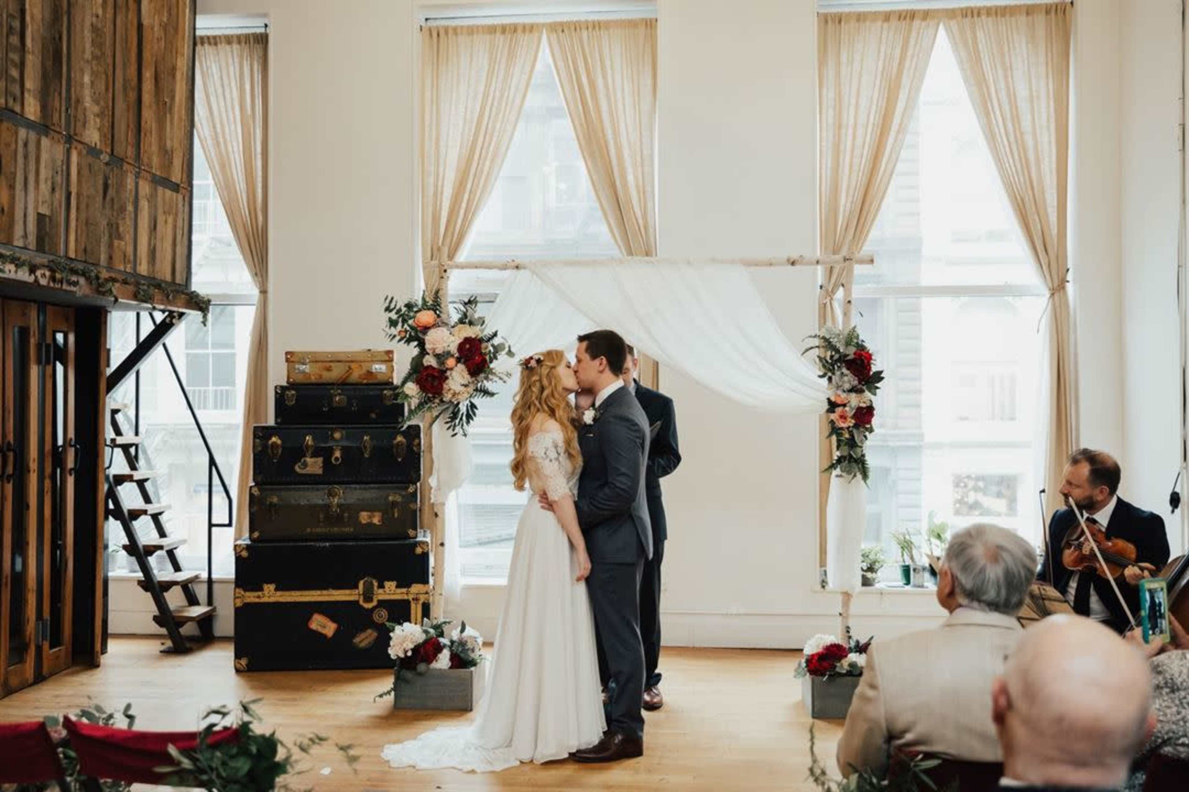 A bride and groom exchange vows under a floral arch in a decorated venue with large windows and vintage luggage in the background.