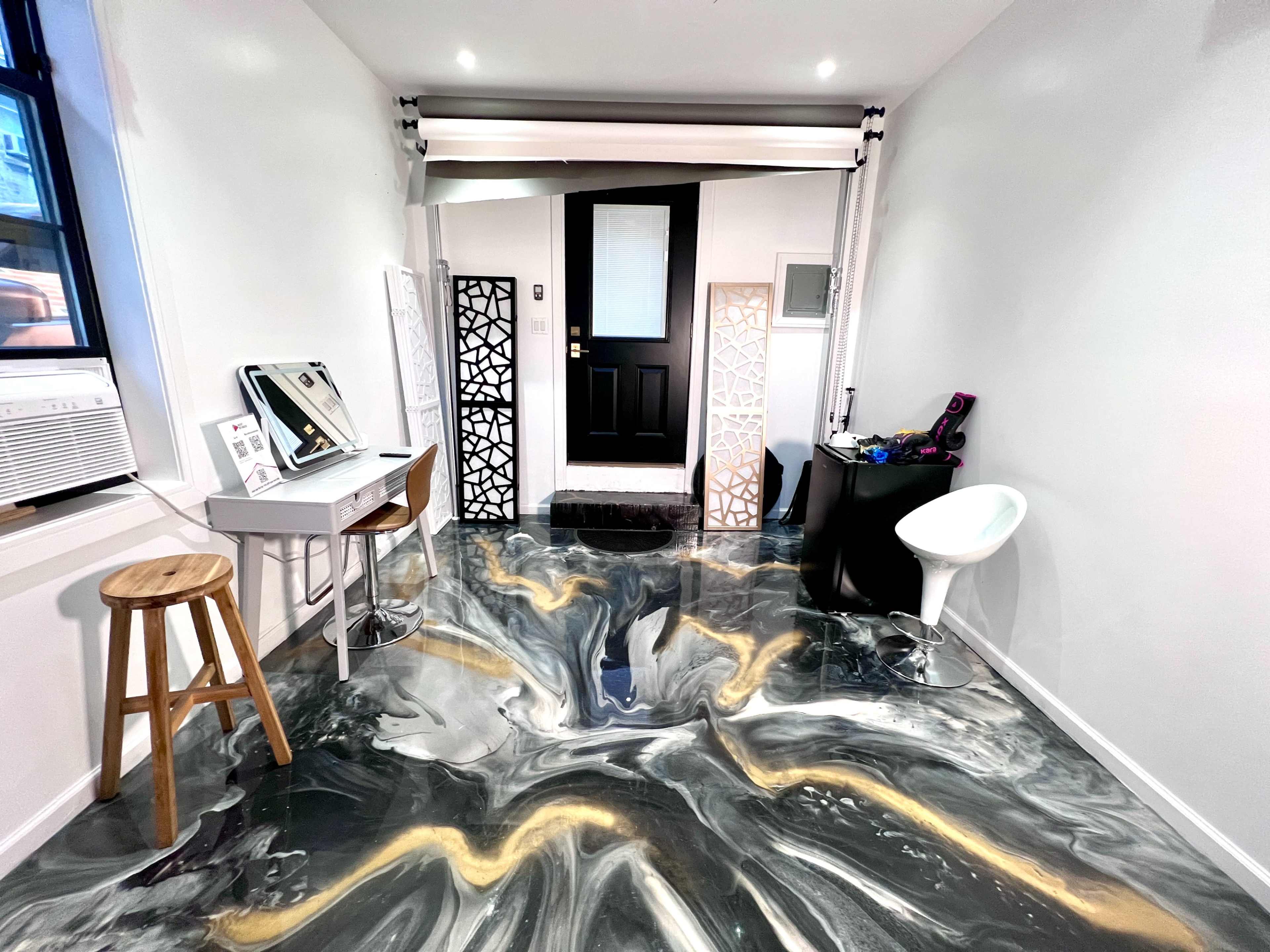 The image shows a modern indoor space featuring a glossy, marbled floor, a small desk with a mirror, a stool, and an entrance with a black door framed by decorative panels.