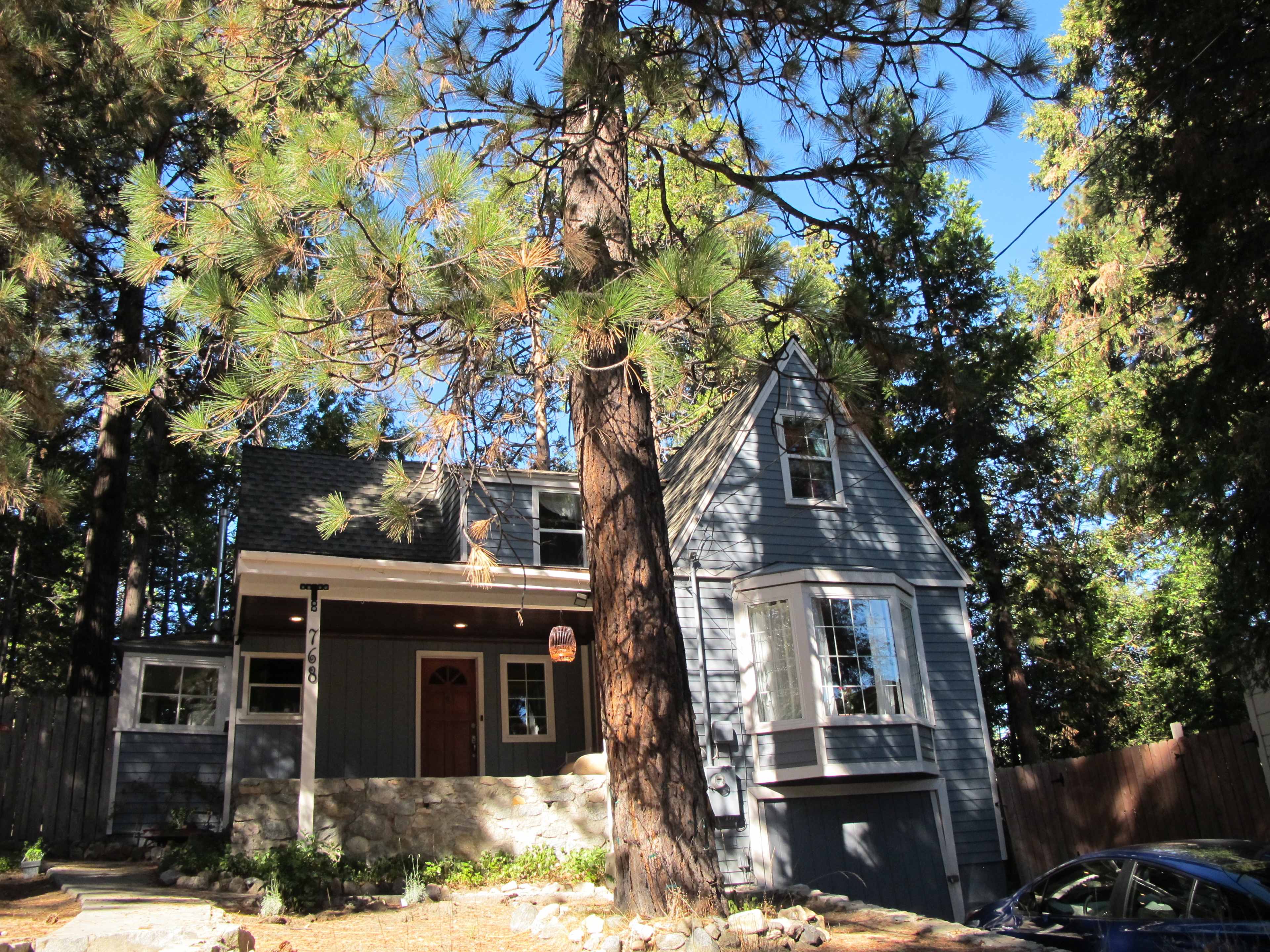 A gray two-story house with a steep roof is situated among tall pine trees.