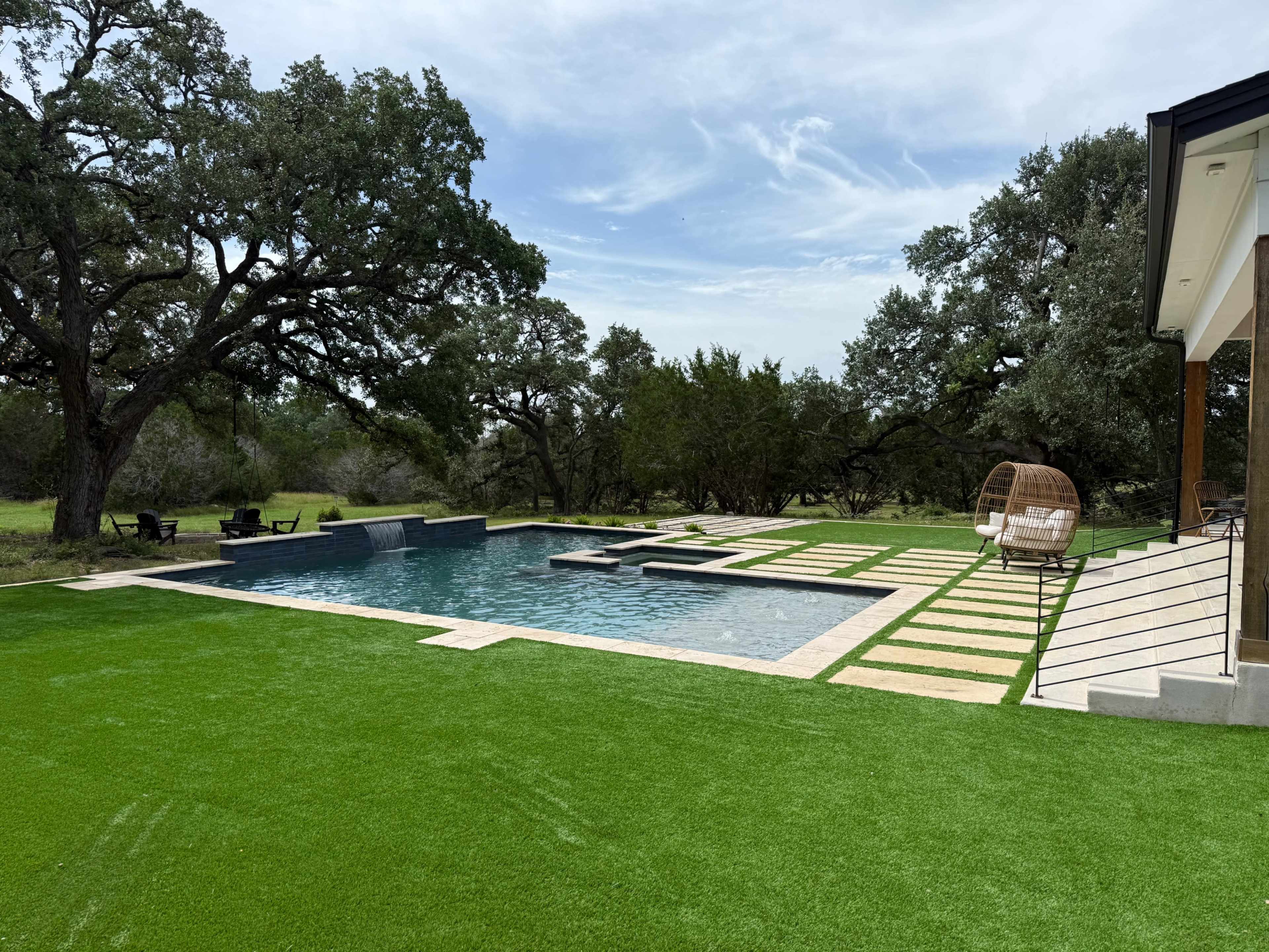 The image shows a modern swimming pool surrounded by artificial grass and strategically placed stone pavers, set against a backdrop of trees.