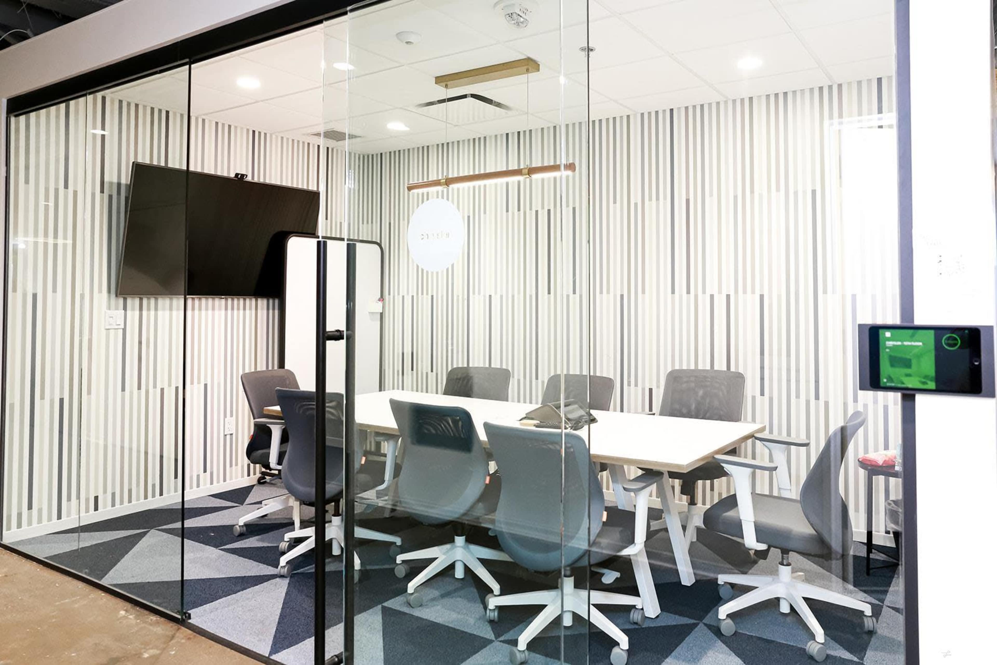 The image shows a modern glass-walled conference room with a white table, gray chairs, and a television mounted on the wall.